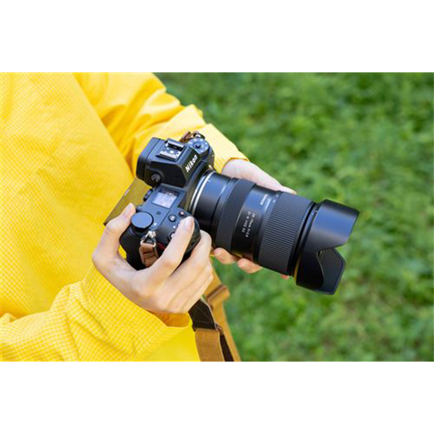 Una persona tiene una fotocamera nera con un obiettivo, indossa una camicia gialla ed erba verde.