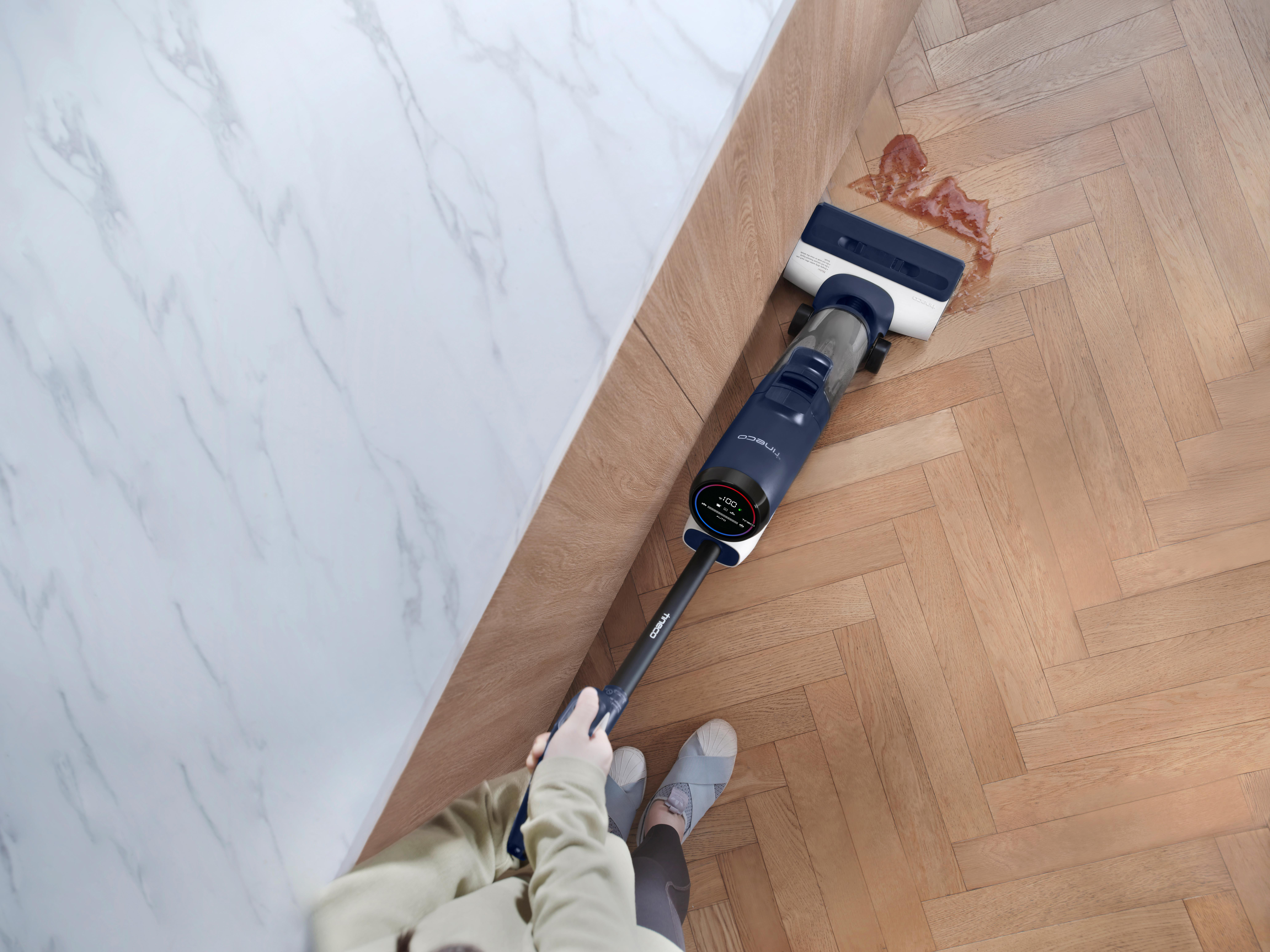 Eine Person, die einen kabellosen Staubsauger auf Holzböden benutzt, um einen roten Fleck in der Nähe einer weißen Wand zu reinigen.