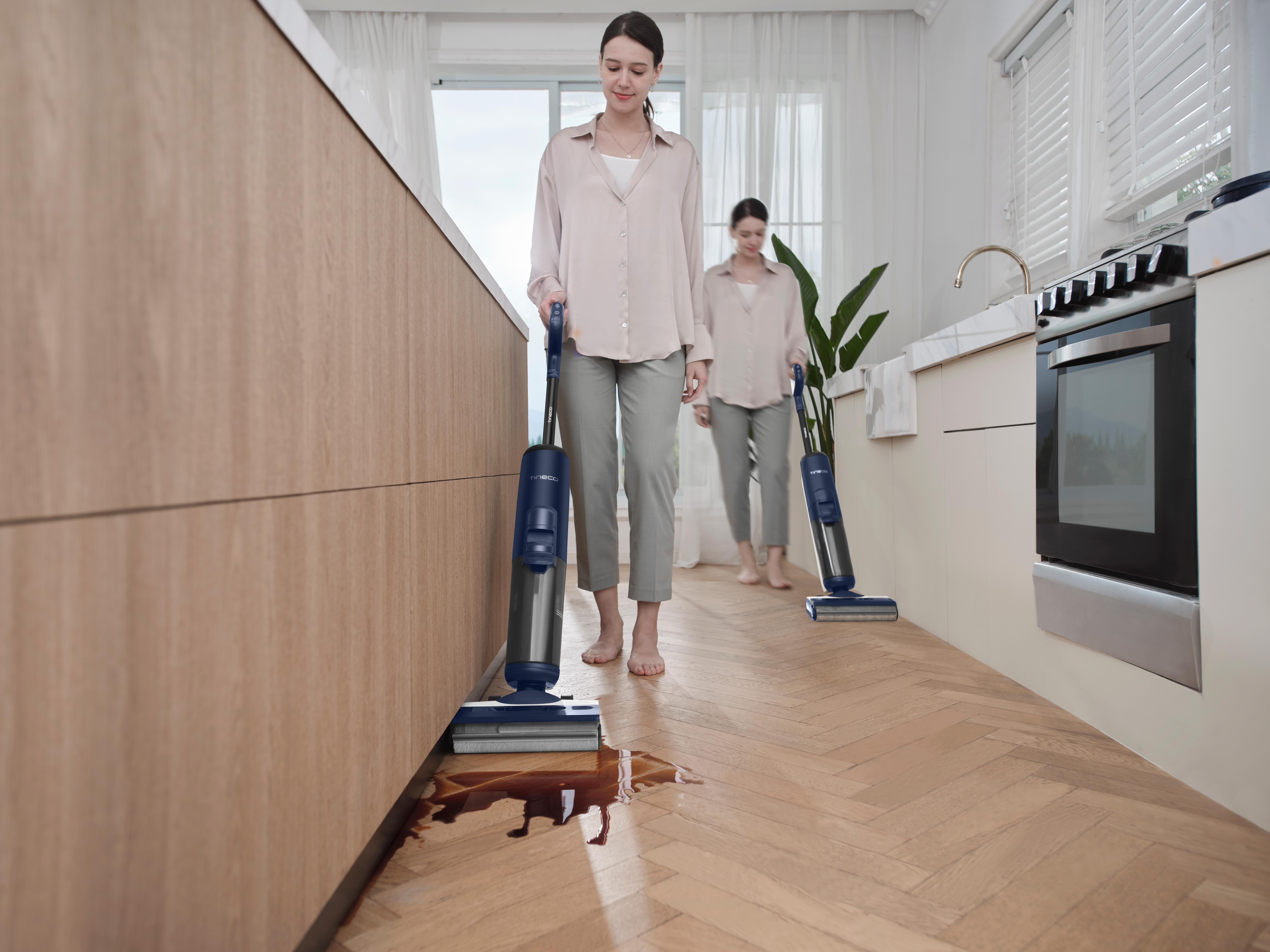 Zwei Frauen reinigen einen Küchenboden mit einem Nass-Trocken-Staubsauger. Ein verschütteter Fleck auf dem Boden ist sichtbar.