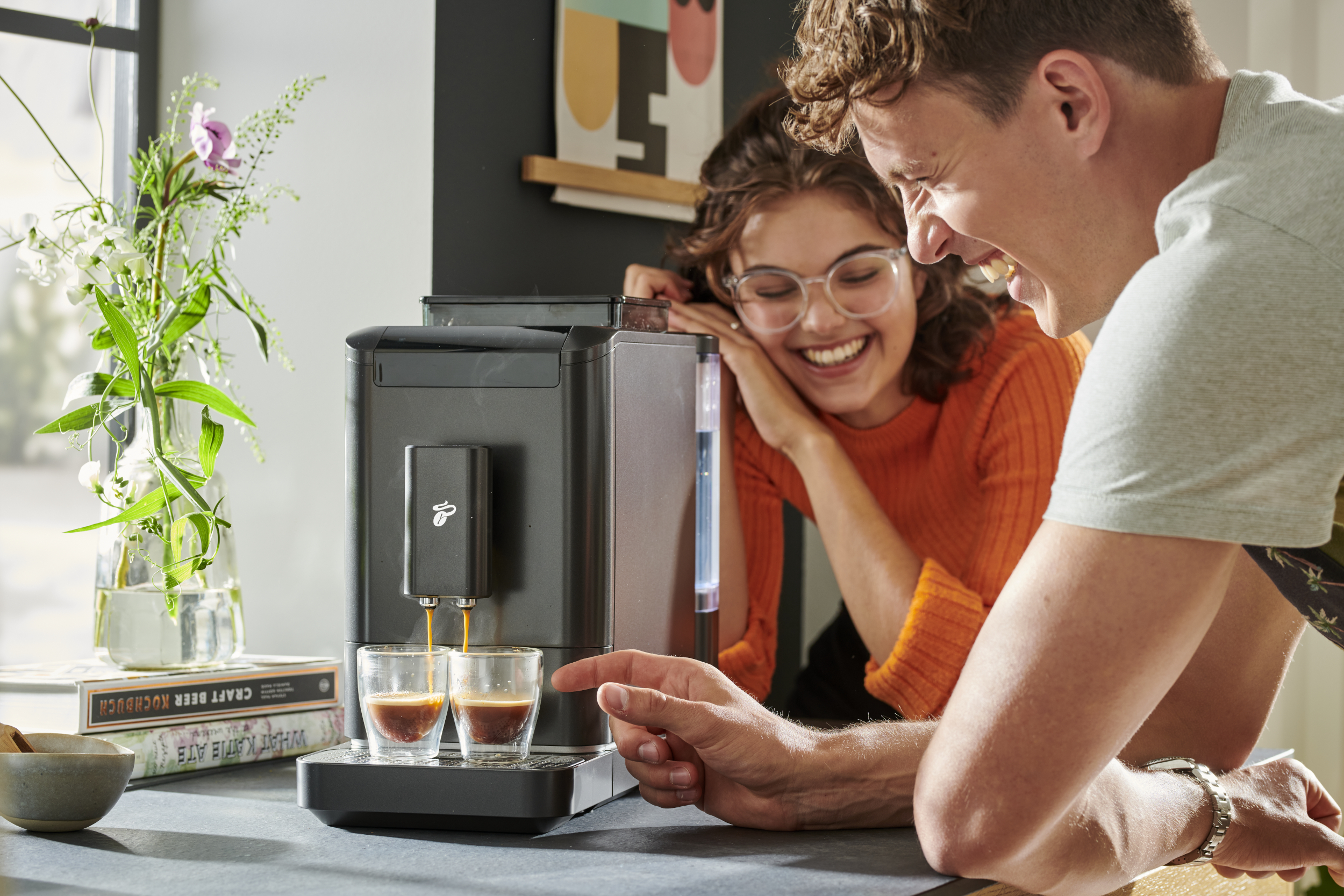 Dwie osoby śmieją się w pobliżu ekspresu do kawy nalewającego kawę do szklanek.