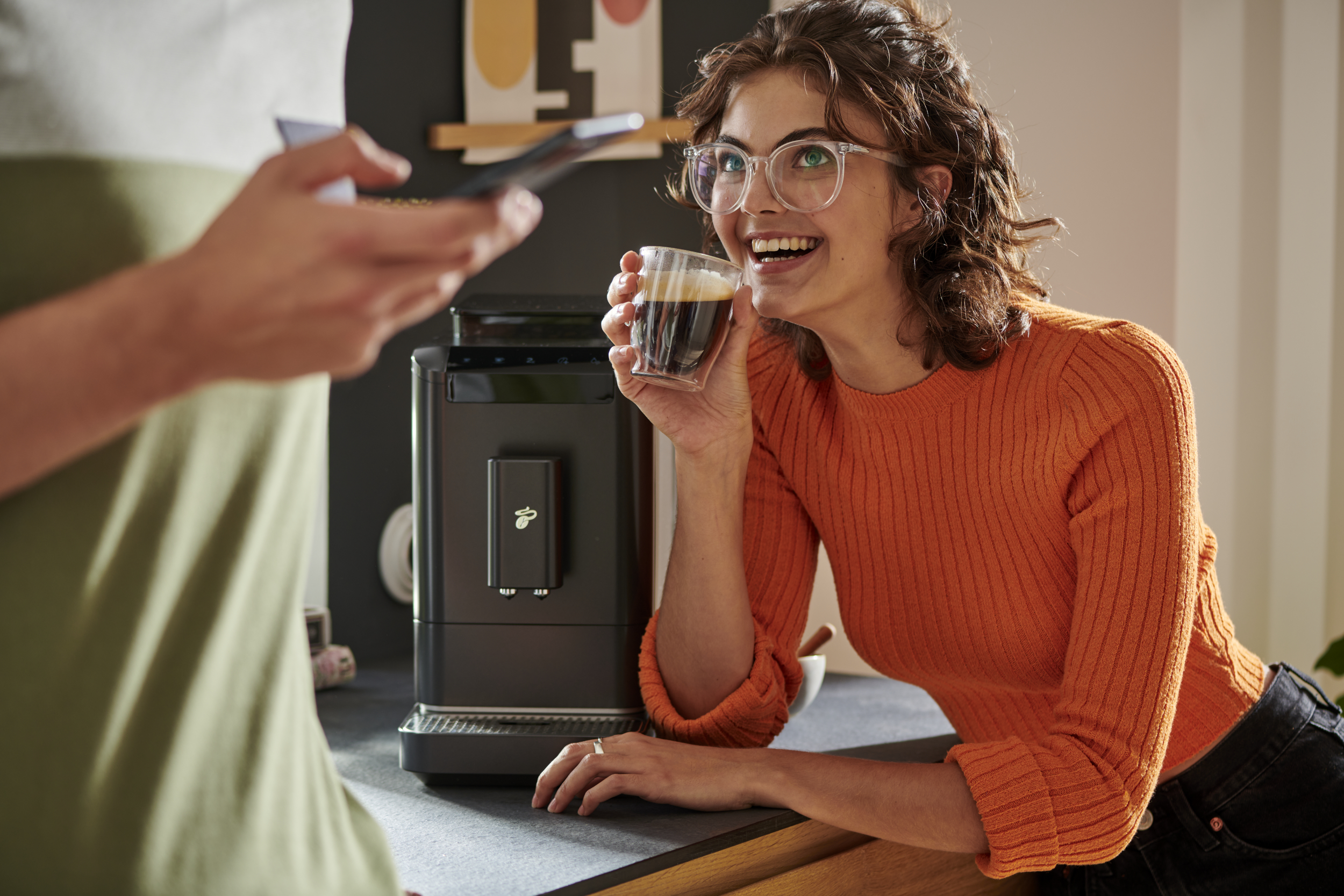 Uśmiechnięta kobieta trzymająca filiżankę kawy, z ekspresem do kawy w pobliżu.