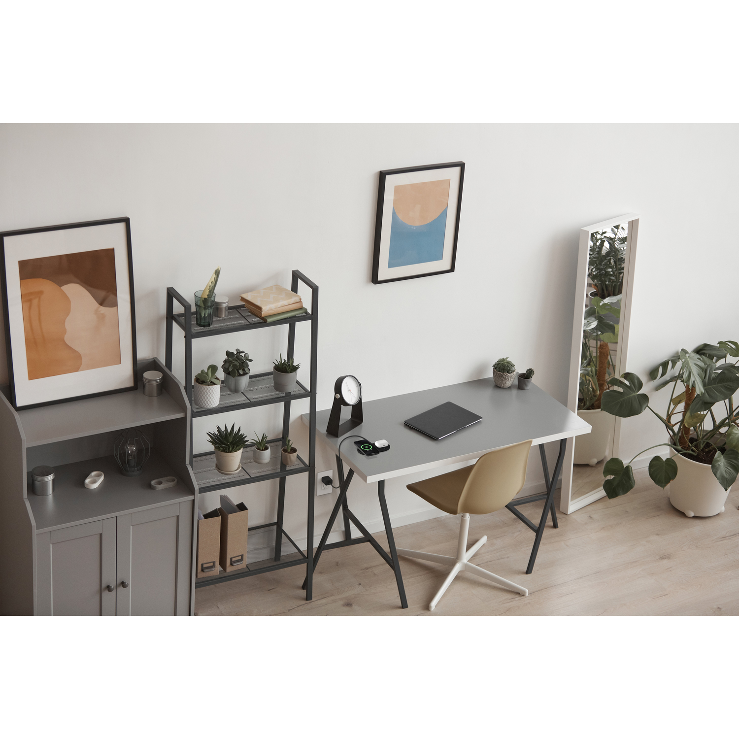 Een kamer met bureau, planken en planten. Een schilderij en een spiegel leunen tegen de muur.