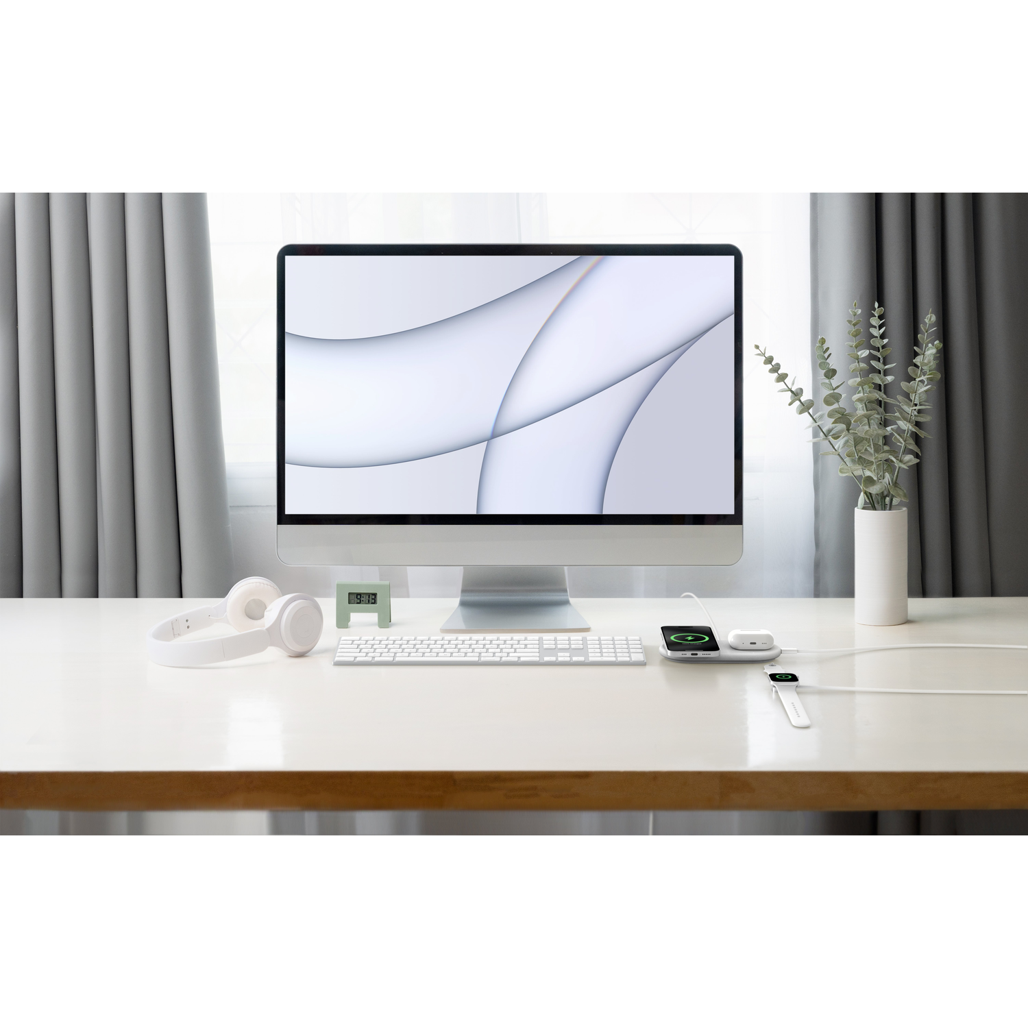 White desk with a computer, headphones, keyboard, clock, phone, and a vase with leaves.