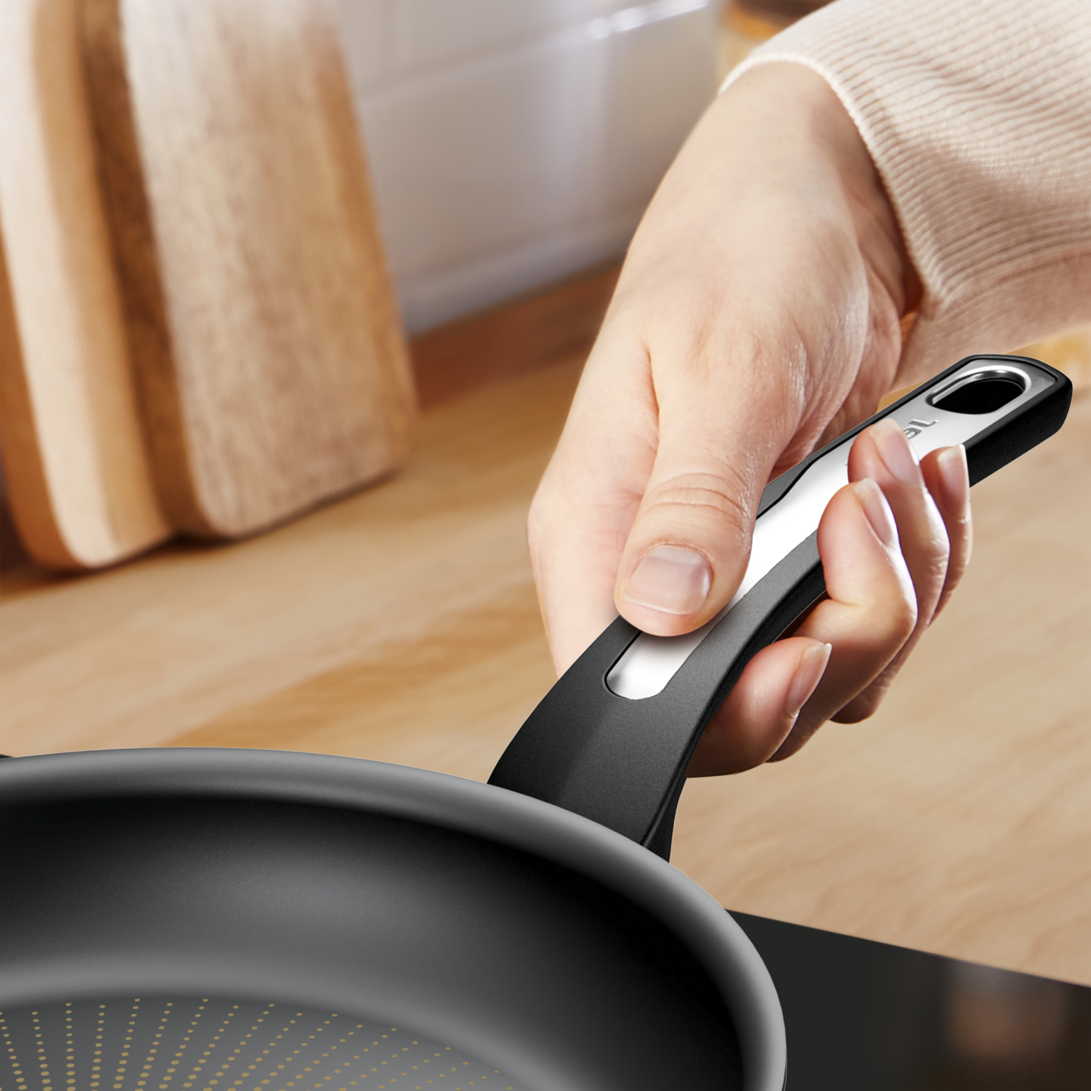 Ręka osoby trzyma czarną patelnię ze srebrnym uchwytem. Deska drewniana w tle.