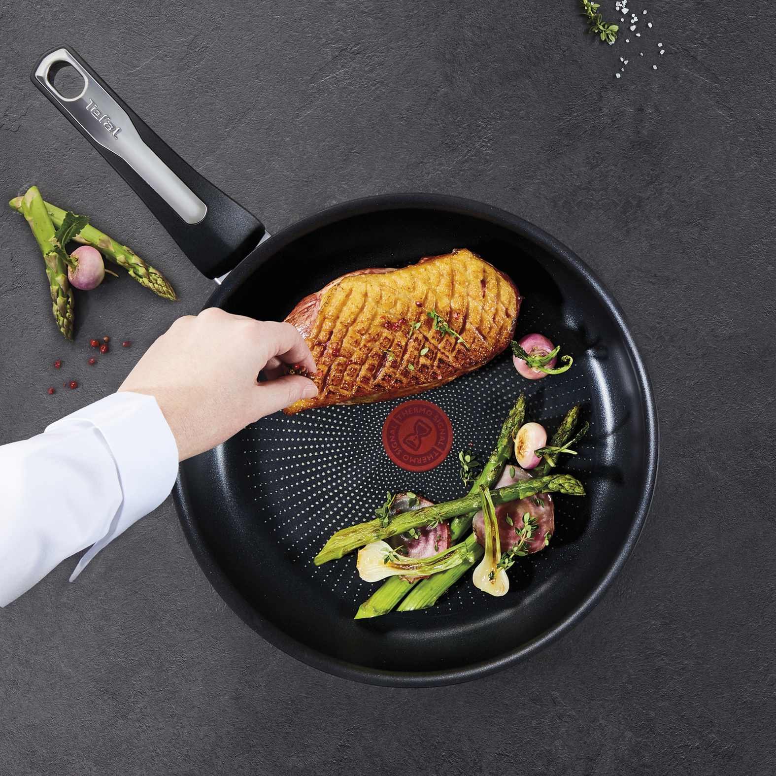 Ręka umieszcza pierś z kaczki na patelni ze szparagami, rzodkiewkami i logo Tefal.