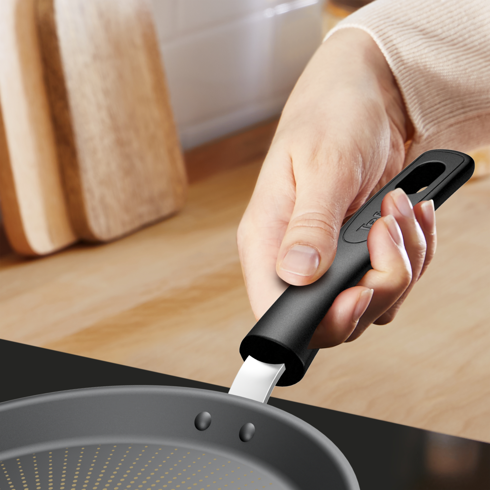 Dłoń trzyma czarny uchwyt patelni nad kuchenką. Drewniane tło.