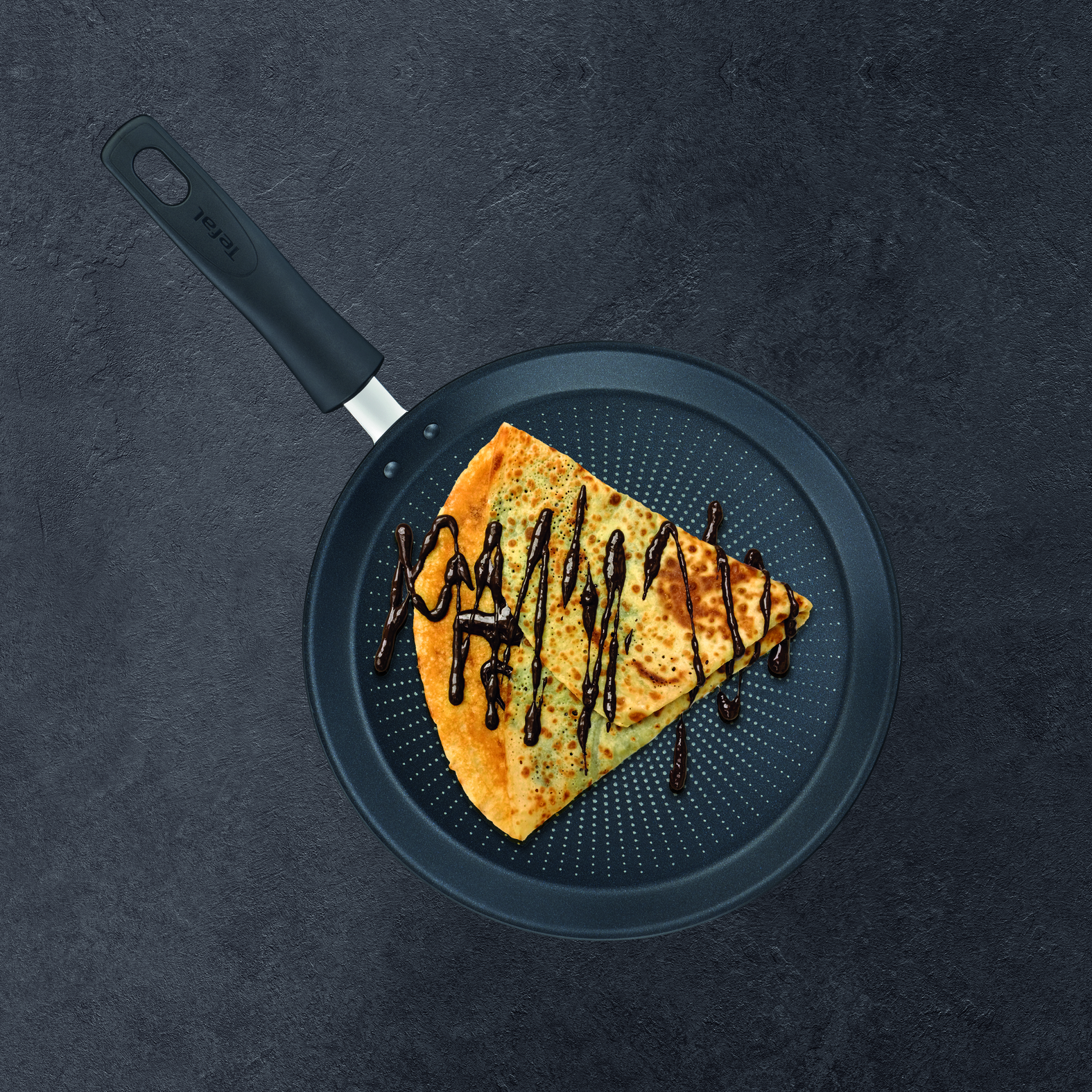 Naleśnik na ciemnej patelni, polany syropem czekoladowym, widok z góry.