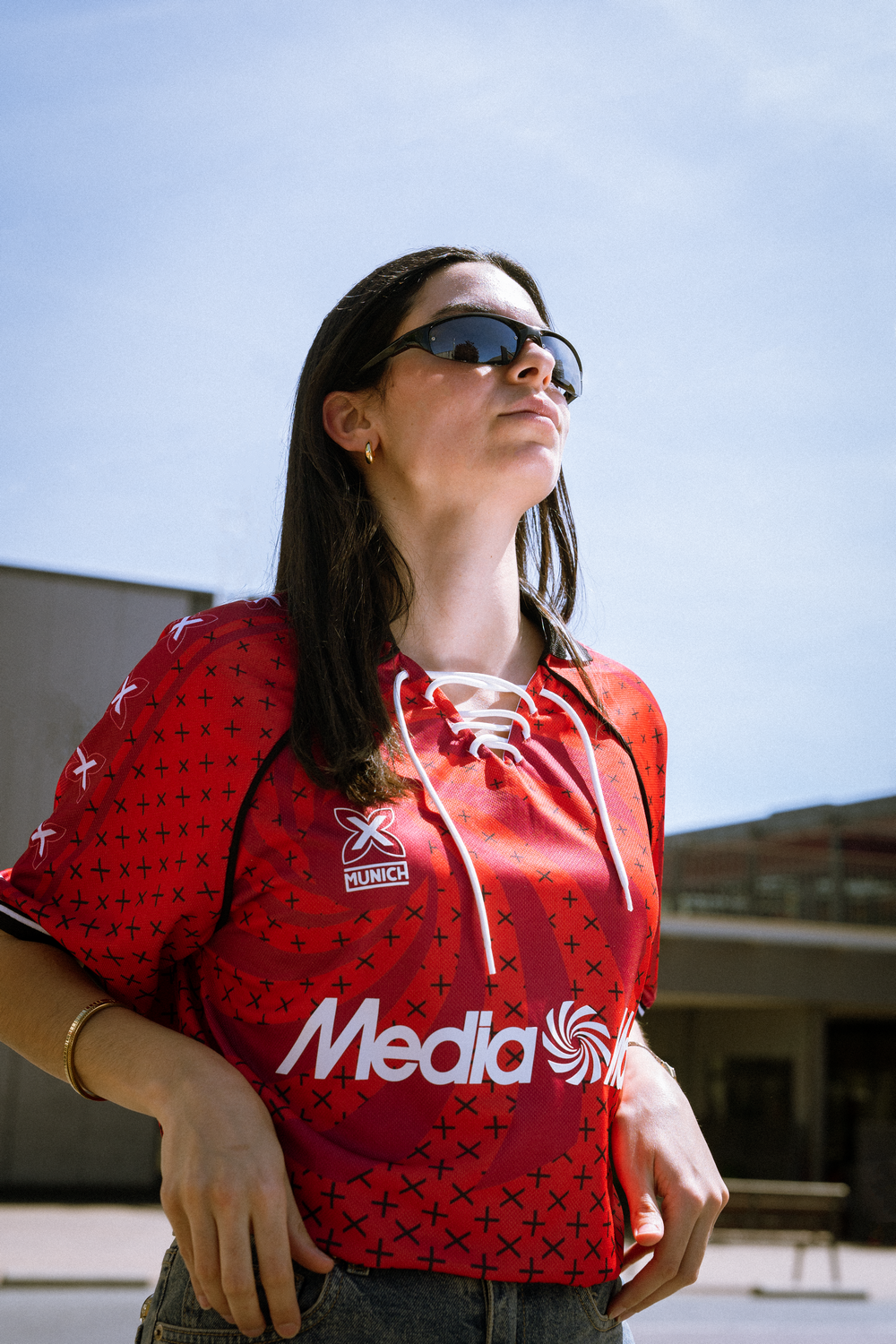 Mujer con camiseta roja y gafas de sol, posando frente a un edificio bajo un cielo azul.