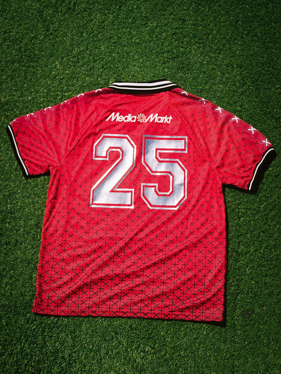 Camiseta de fútbol roja. Número 25 en la espalda. Logo MediaMarkt. Fondo césped verde. Patrón de cruces negras.