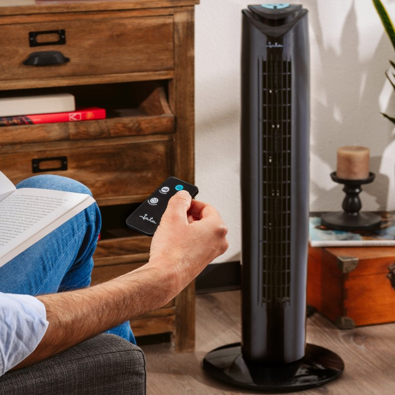 Una persona sostiene un control remoto para un ventilador de torre negro en una habitación con muebles de madera.