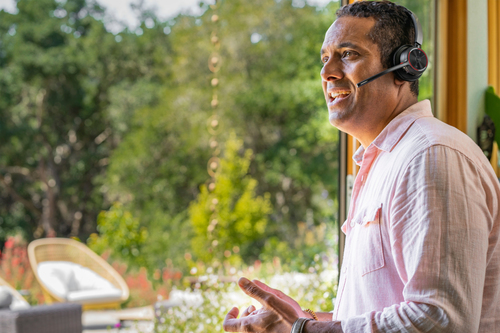 Hombre con auriculares, hablando. Camisa rosa, tono de piel claro. Ventana y vista exterior. Día soleado.