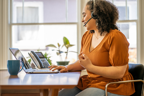 Mujer en escritorio usando laptop, headset, hablando. Taza azul, teléfono y planta en el fondo.
