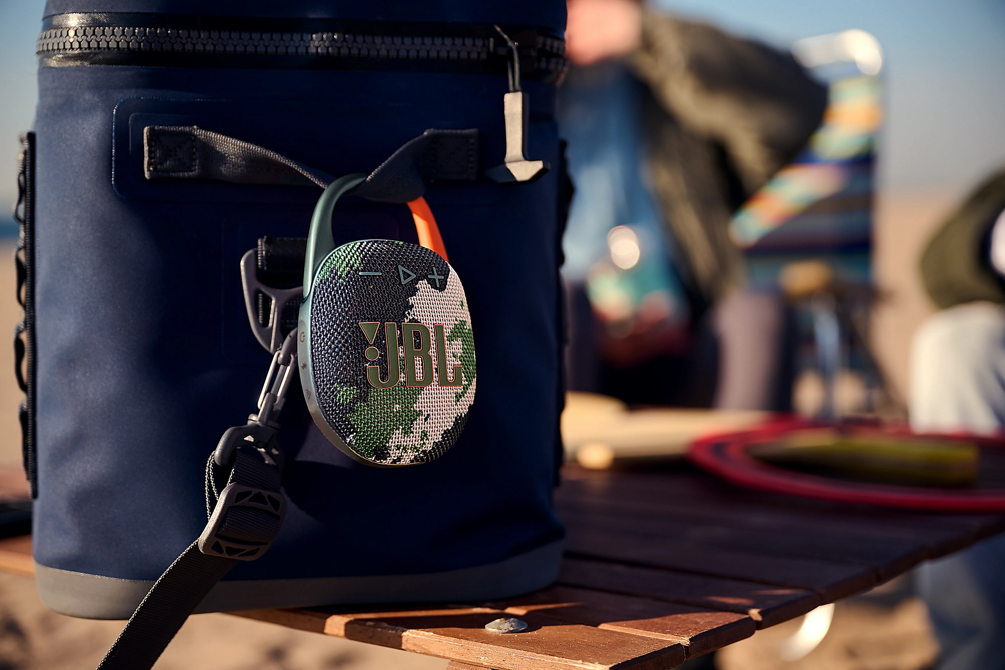 Głośnik JBL przymocowany do granatowej chłodziarki na drewnianym stole w pobliżu plaży.