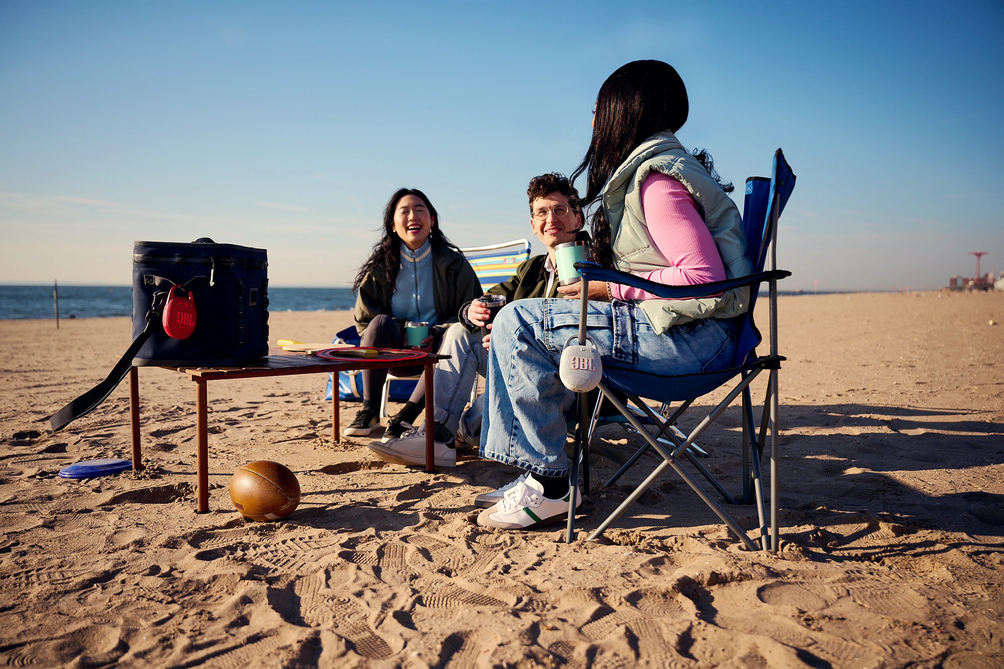 Trzy osoby siedzące na plaży, cieszące się piknikiem z lodówką, głośnikami i piłką nożną. Słoneczny dzień.