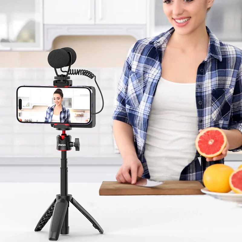Nő telefonnal filmez állványon, grapefruitot tart a konyhában.