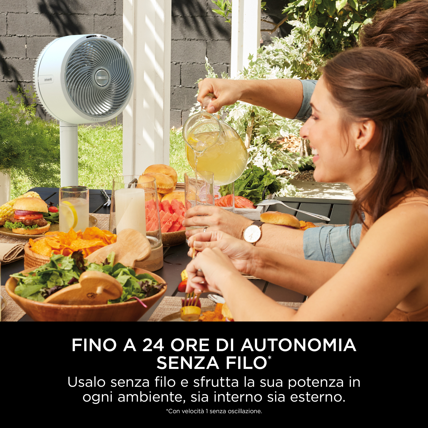Persone a tavola all'aperto con cibo e un ventilatore. Una persona sta versando una bevanda, testo sotto.