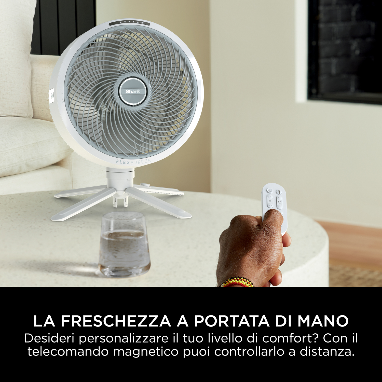 Ventilatore bianco e grigio con mano che tiene il telecomando. Bicchiere d'acqua.
