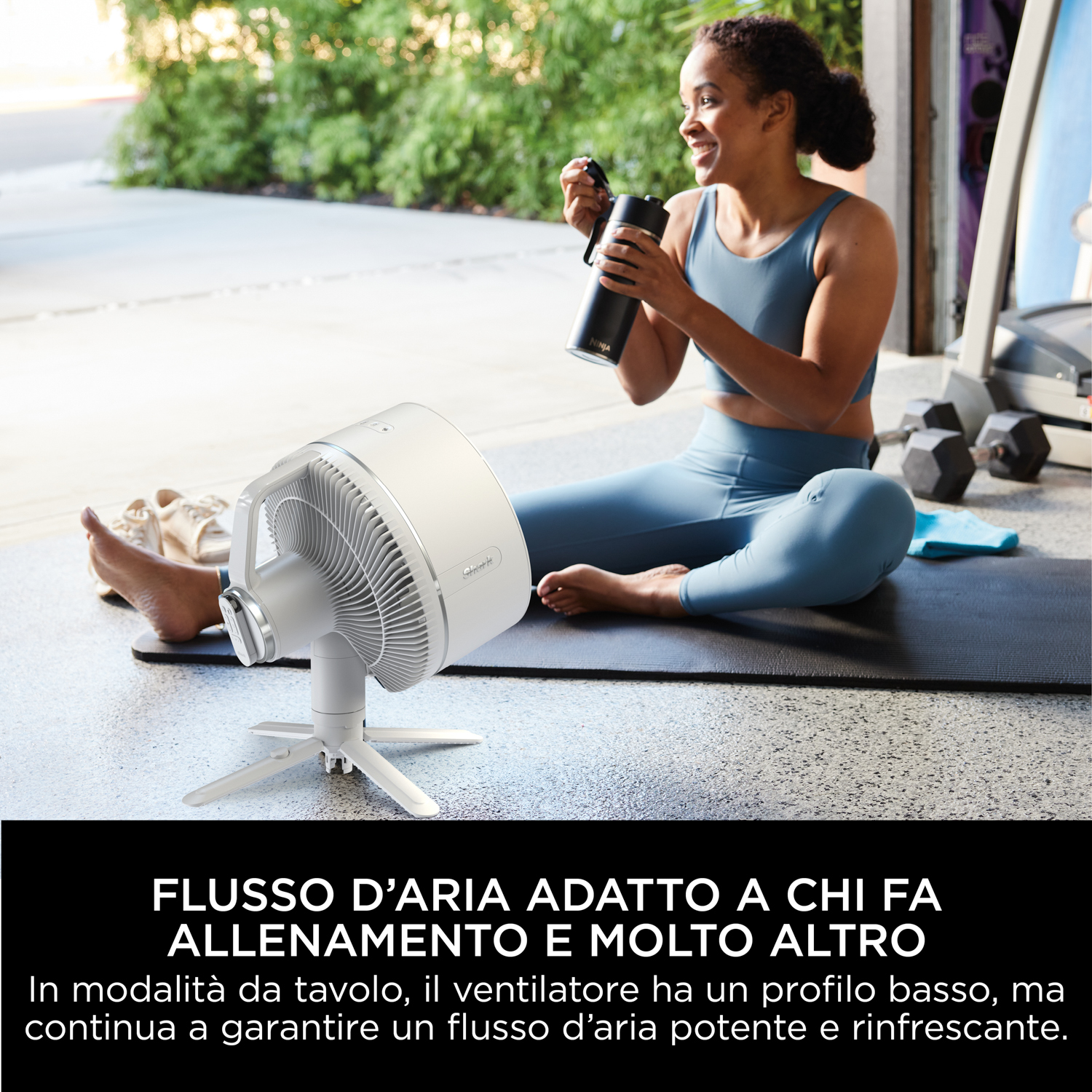 Donna seduta vicino a un ventilatore bianco, con una bottiglia in mano. Sfondo bianco.
