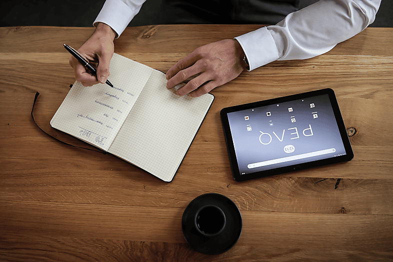 Egy személy kezei egy noteszbe írnak, és egy tablet a 'peaq' szóval egy faasztalon.