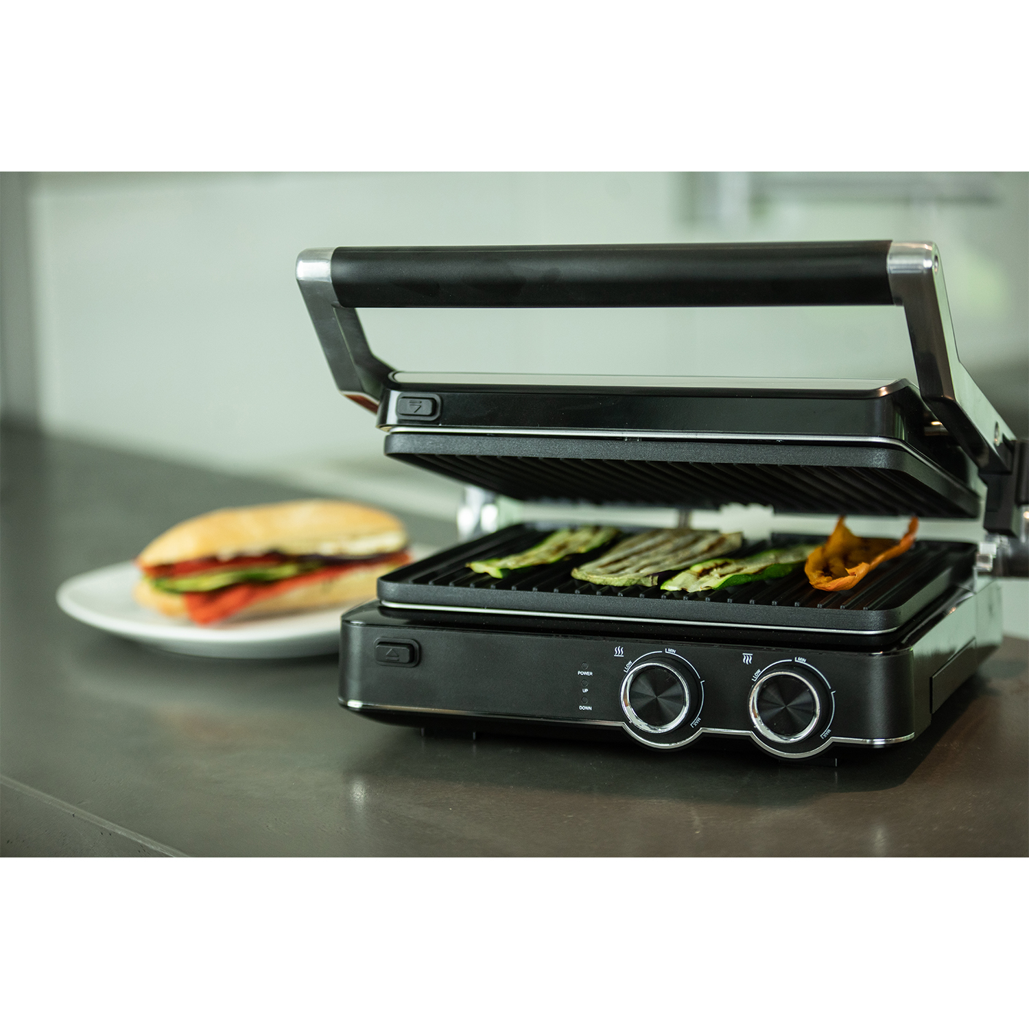 Griglia nera aperta con verdure che cuociono all'interno, un panino e comandi. Su un piano cucina.