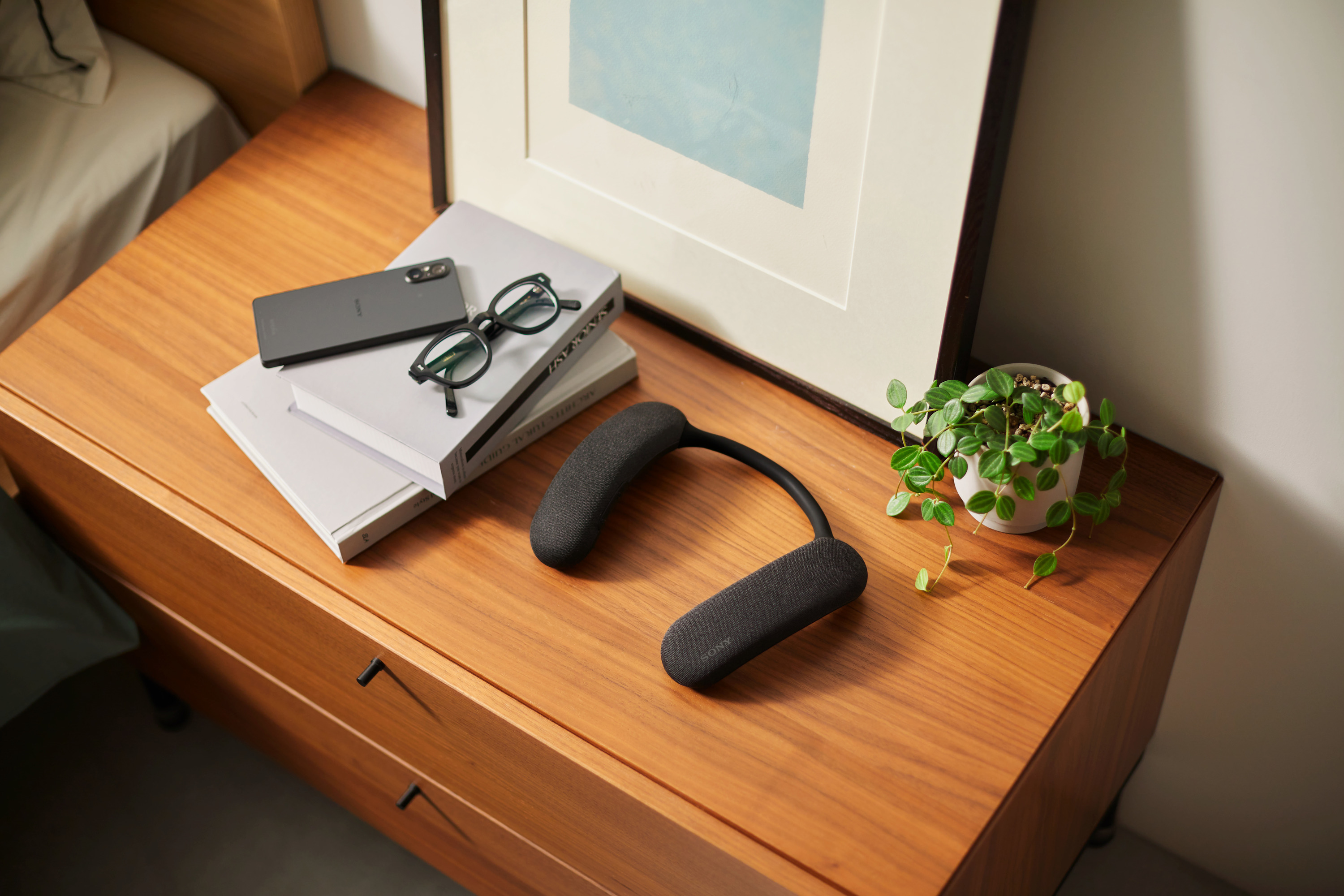 Un bureau avec un smartphone, des lunettes, des livres, une plante et une enceinte de cou dessus.