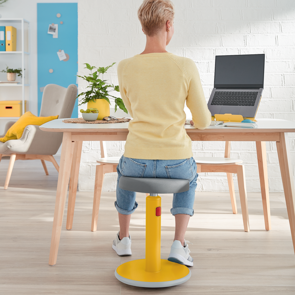 Nő laptopot használ az asztalnál, sárga-szürke ergonomikus széken ül.