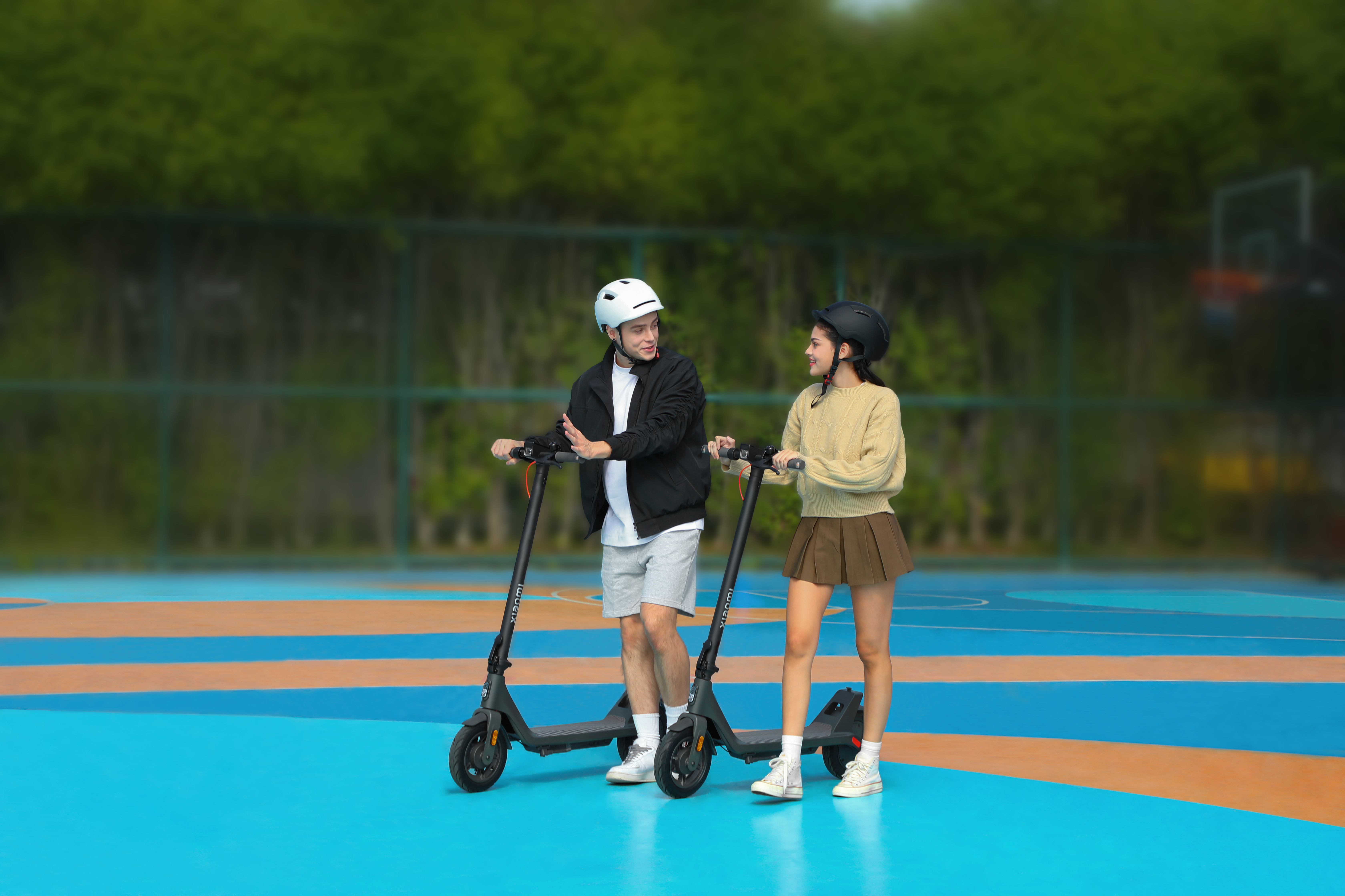 Zwei Personen mit Rollern auf einer blau-weißen Oberfläche, mit grünen Bäumen im Hintergrund.