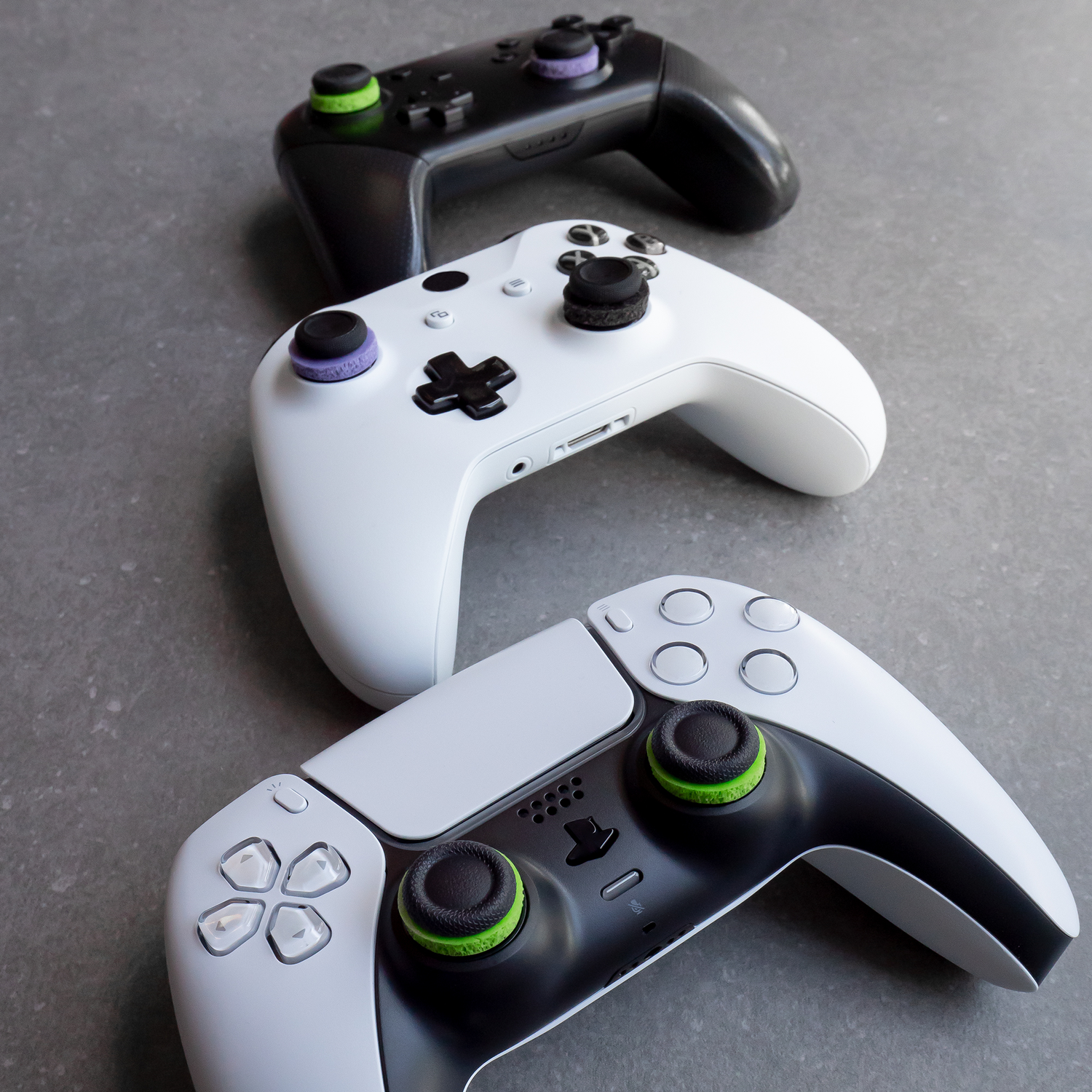 Three game controllers, black, white, and white with black. Each has colored thumbsticks.