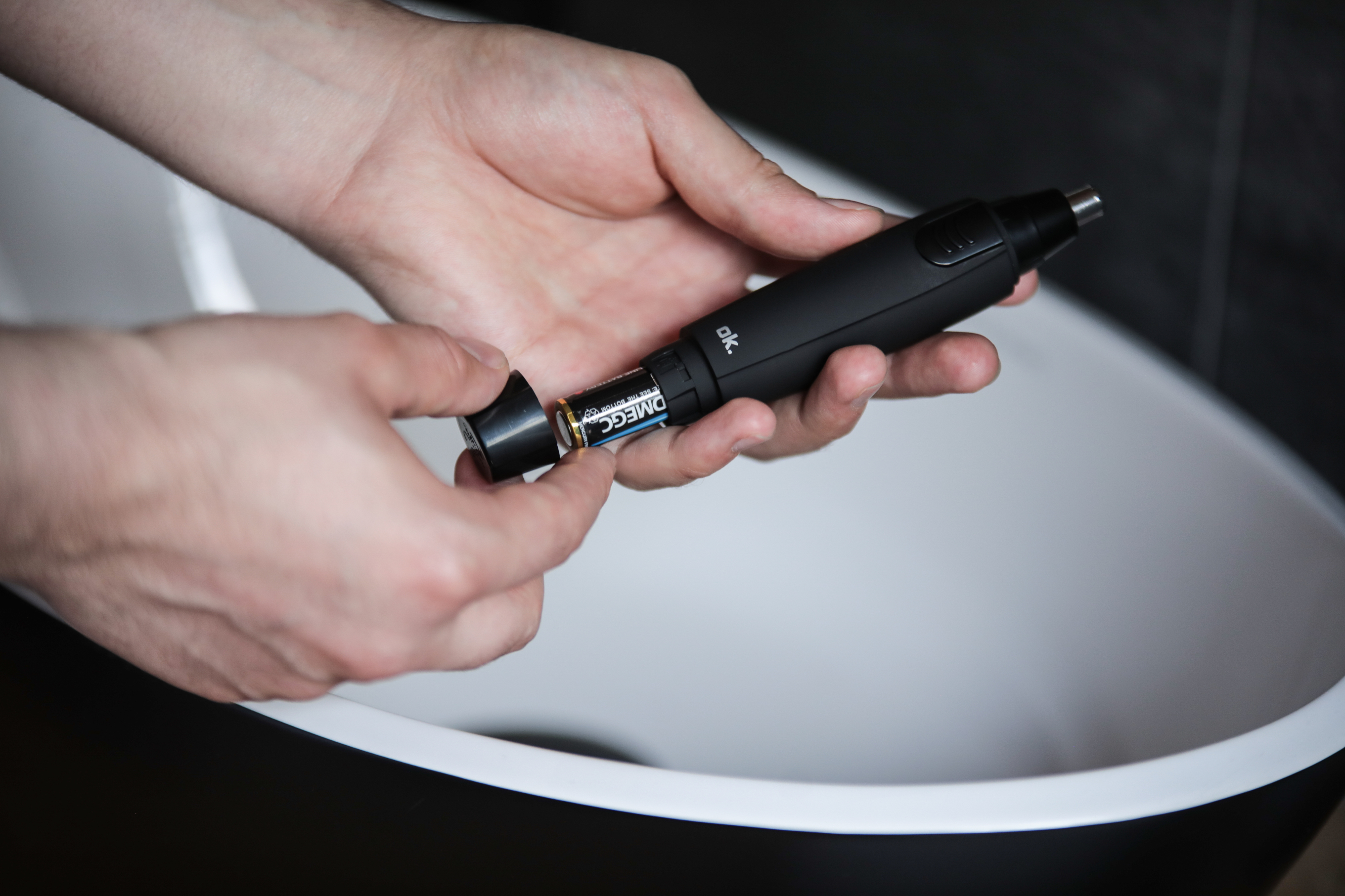 Dłonie otwierające czarny trymer do włosów w nosie, aby włożyć baterię, nad zlewem.