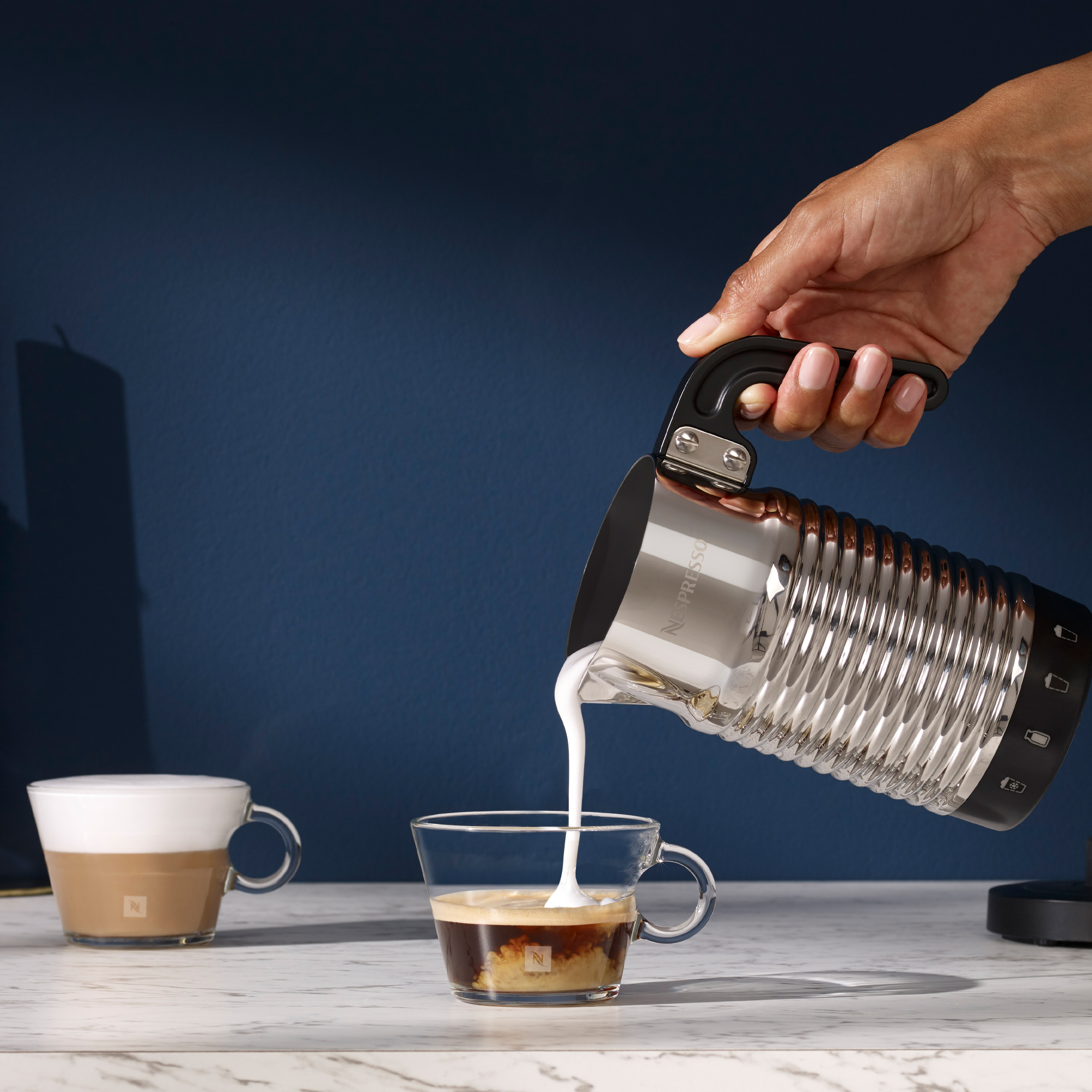 Eine Hand gießt Milch in eine Glastasse mit Kaffee, in der Nähe einer Kaffeemaschine und Tasse.