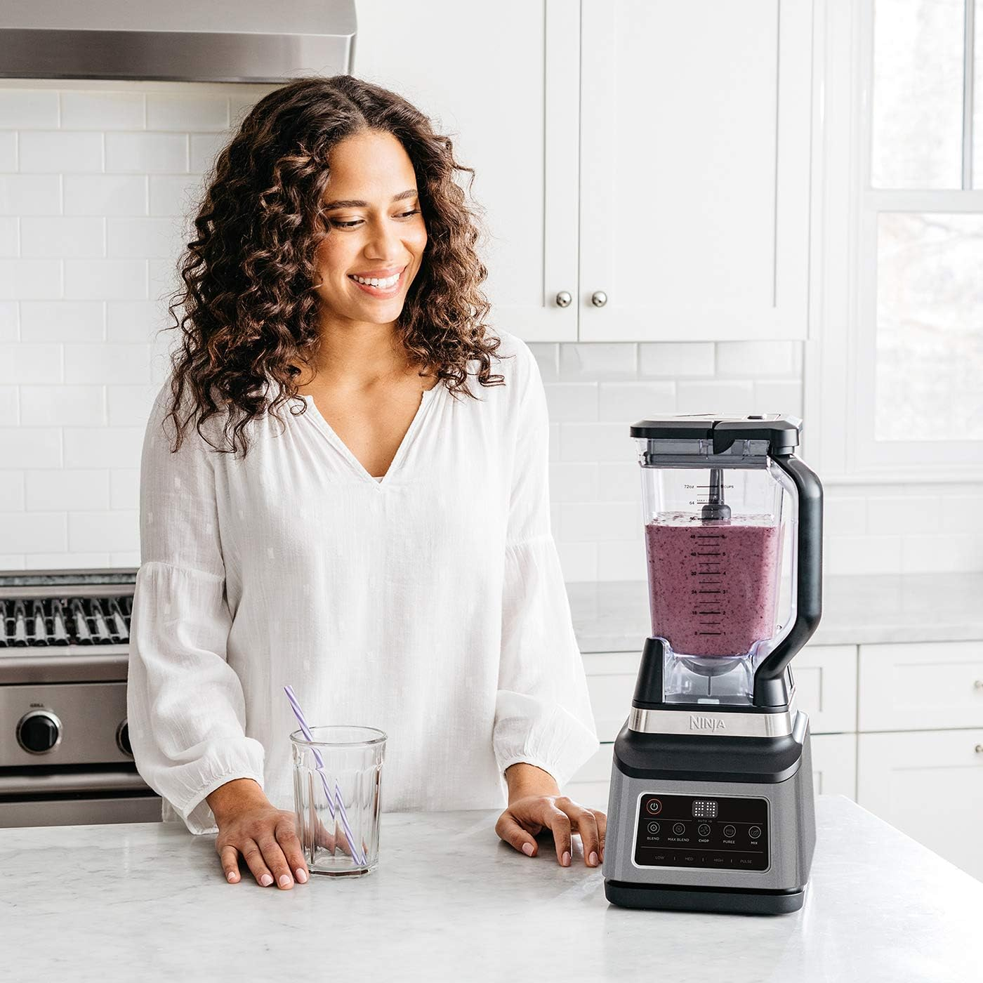 Femme utilise un blender, verre et blender. Fond de cuisine blanc, souriante.