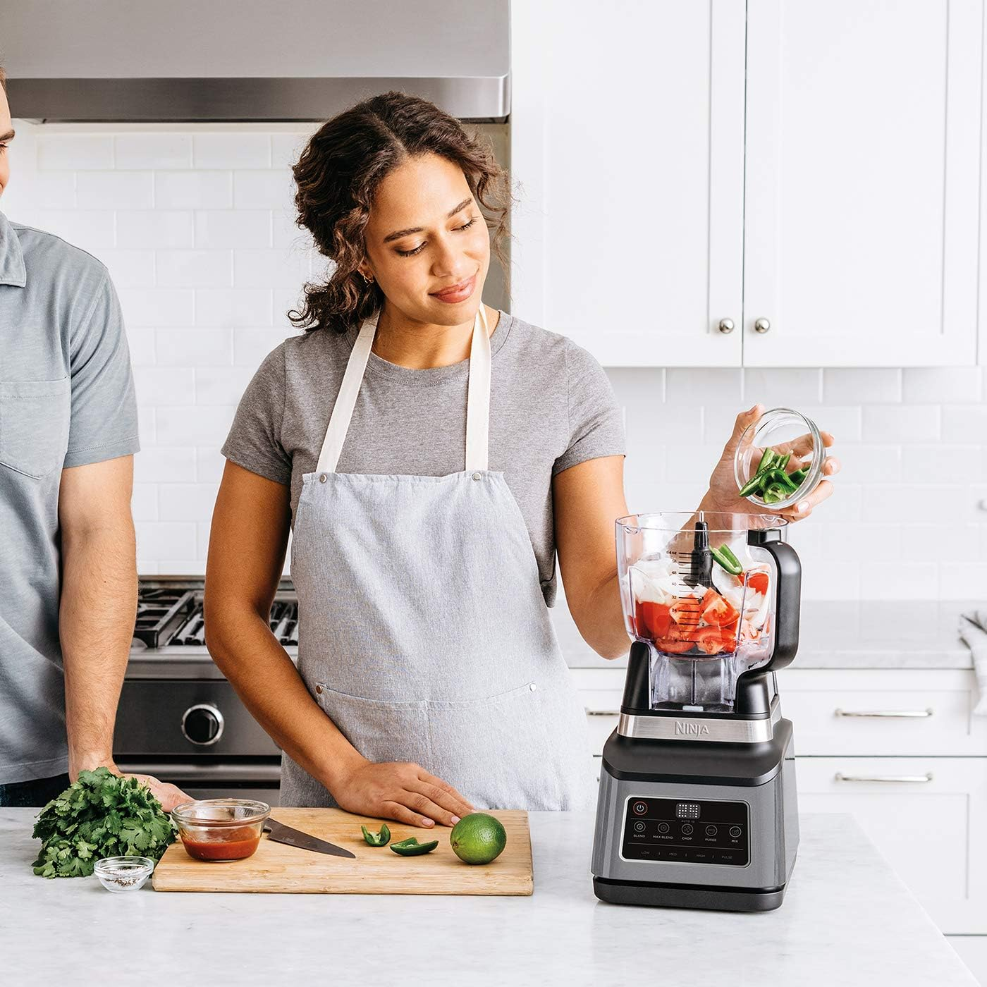 Une femme ajoute des poivrons verts dans un mixeur avec des tomates et des oignons. Un homme se tient à côté d'elle. Ingrédients sur une planche.