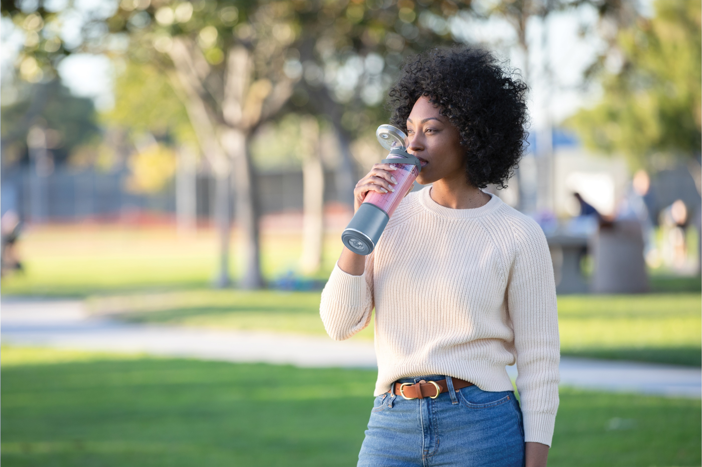 Eine Frau trinkt aus einer Mixerflasche in einem Park. Sie trägt einen Pullover und Jeans, mit grünem Hintergrund.