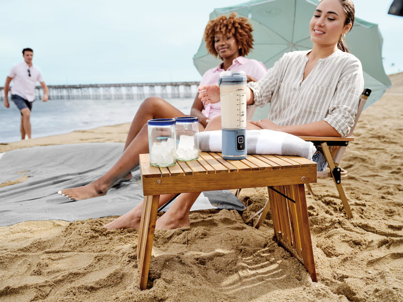 Strandszene: Zwei Frauen mit Mixer auf Tisch, Personen im Hintergrund.