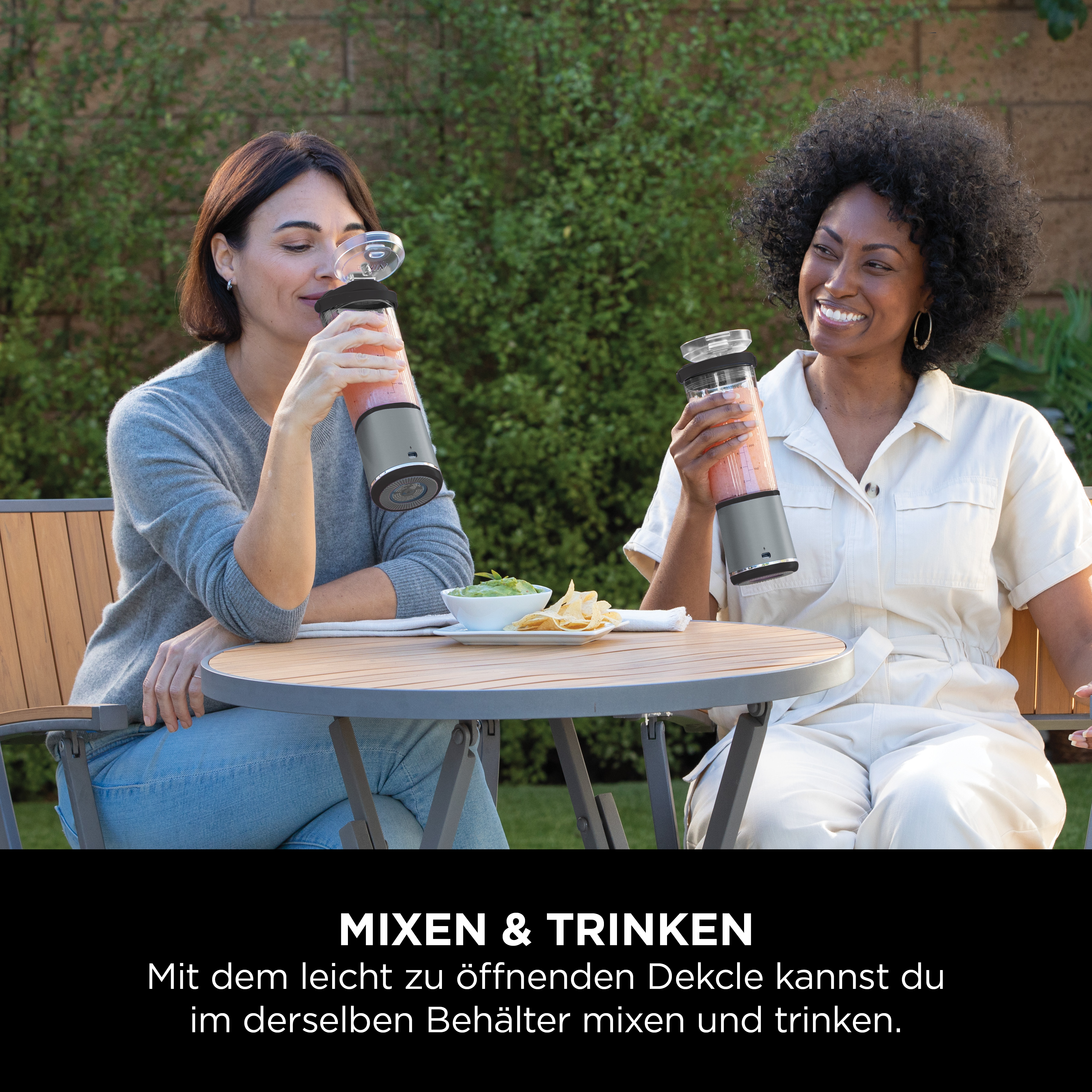 Zwei Personen trinken draußen aus Ninja-Mixern, ein Tisch mit Snacks in der Nähe.