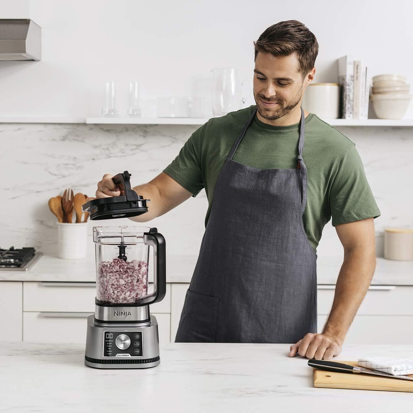 Homme utilisant un robot culinaire Ninja dans une cuisine, préparant de la nourriture. Il porte un tablier.