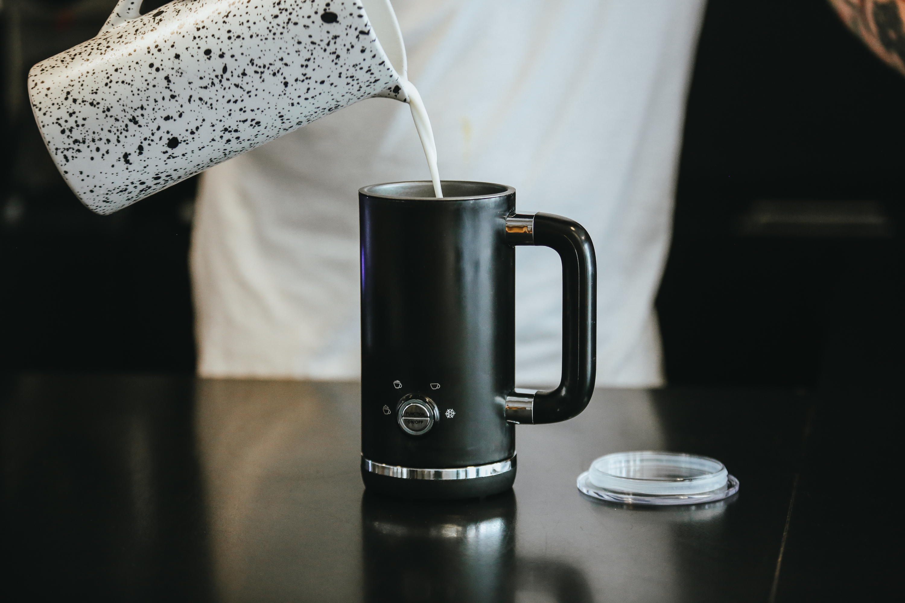 Osoba wlewa mleko do czarnego kubka. Widoczny czarny stół i pokrywka.
