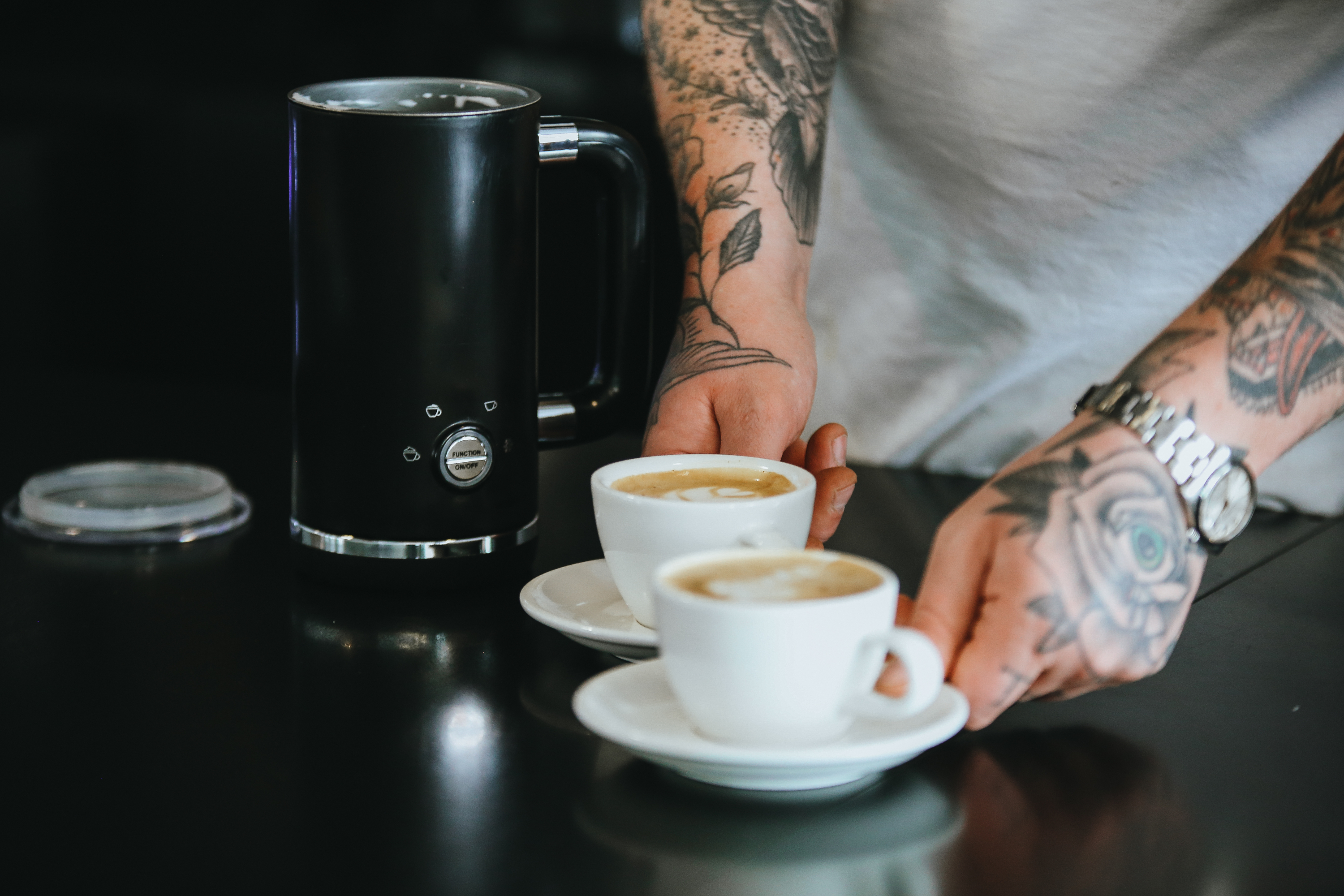 Osoba z tatuażami stawia dwie filiżanki kawy na ciemnym stole obok spieniacza do mleka.