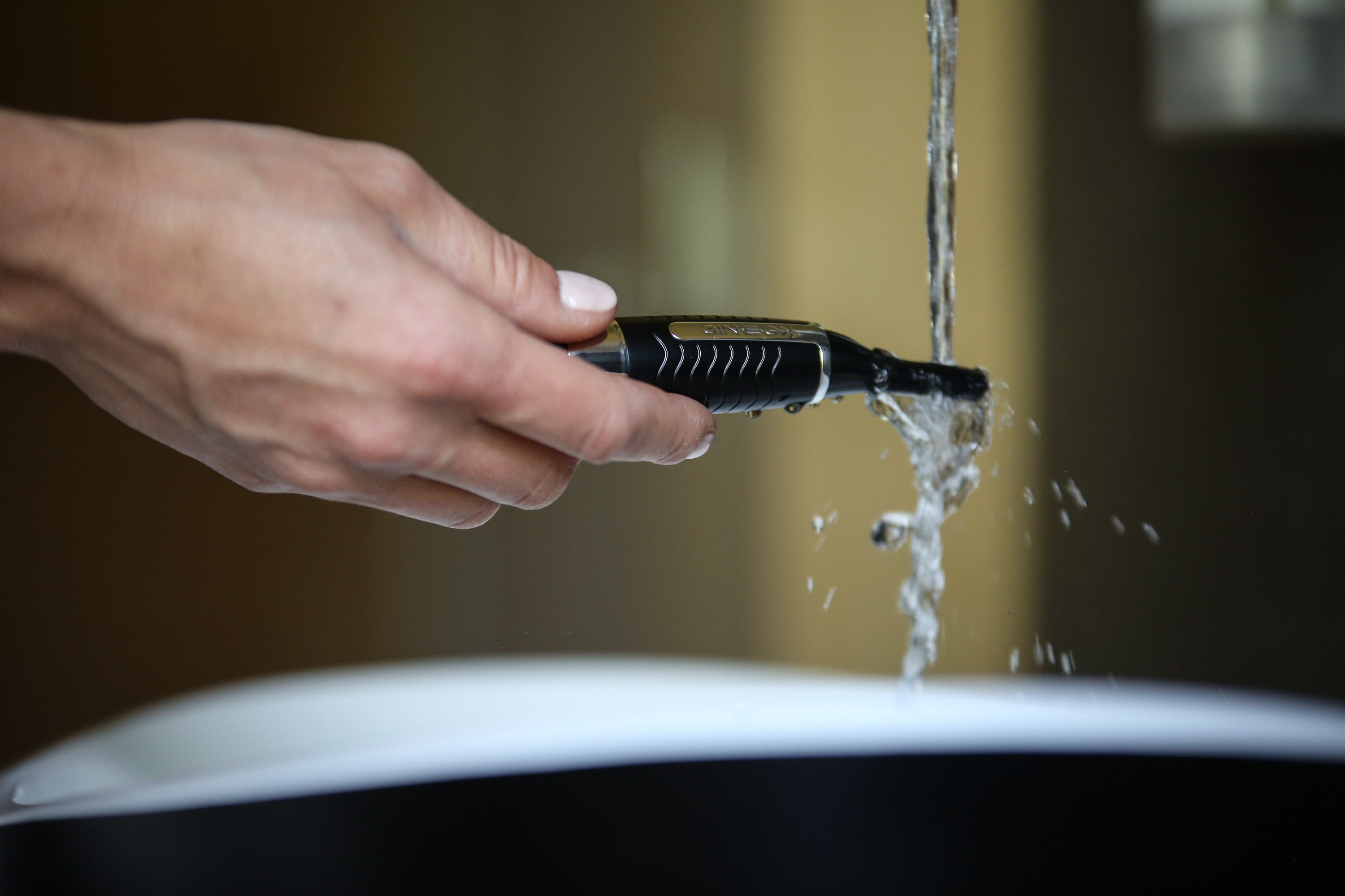 Dłoń trzymająca czarny i złoty trymer pod bieżącą wodą przed zlewem.