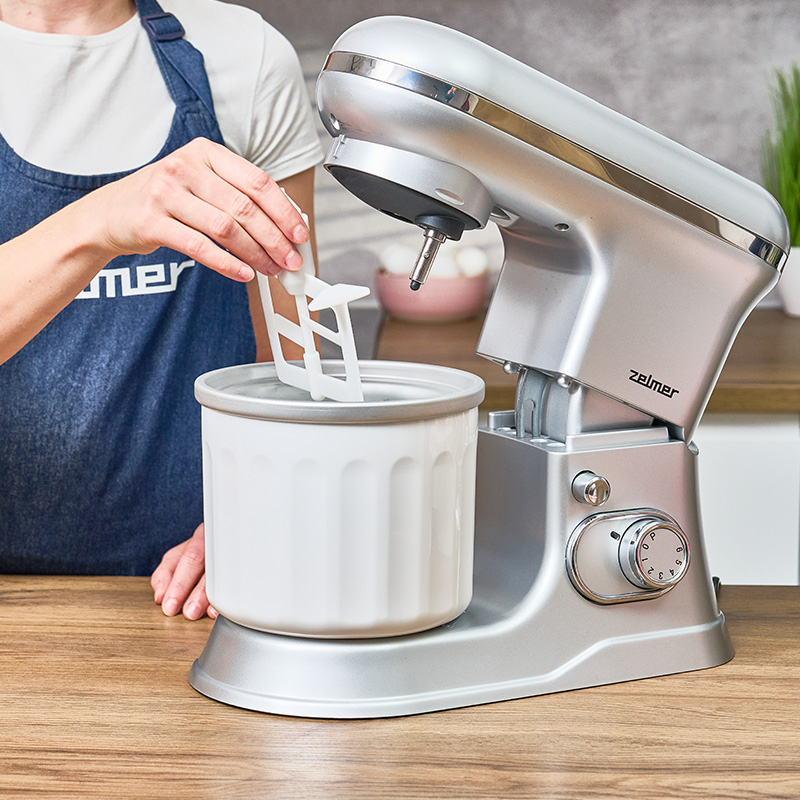 Kobieta w niebieskim fartuchu używa srebrnego miksera. Mikser ma białą misę. W misie znajduje się biała łyżka.