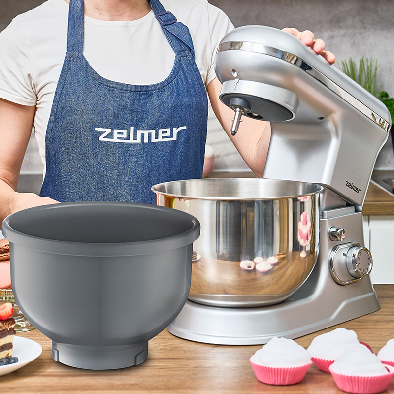 Kobieta używa srebrnego miksera. Misa na stole. Babeczki po prawej. Fartuch z logo.