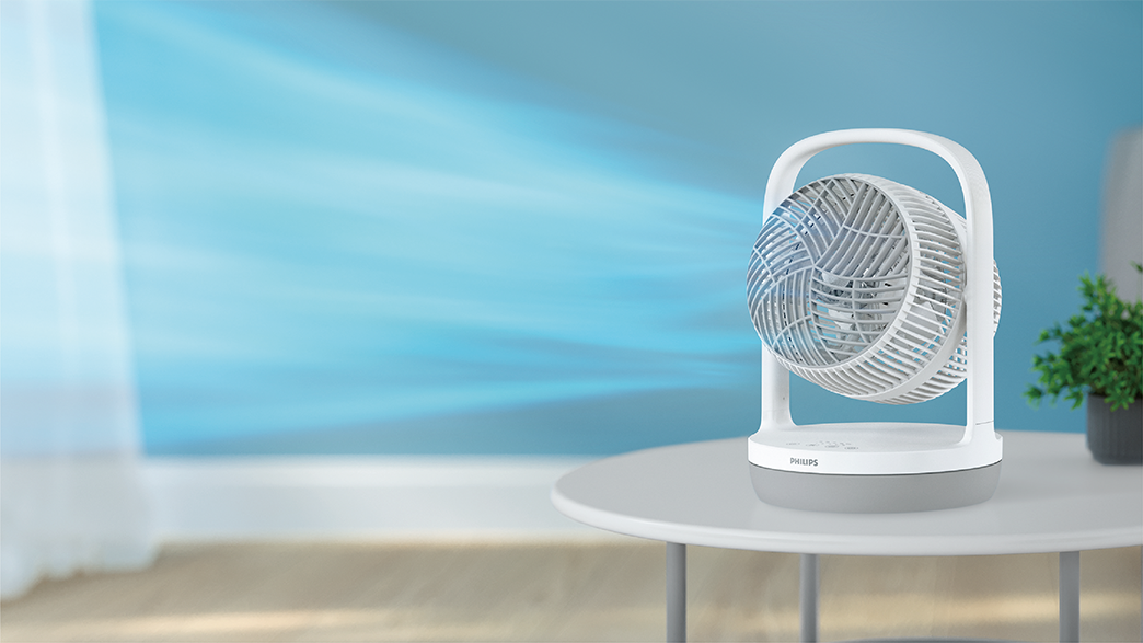 Fehér hordozható ventilátor egy asztalon. A háttér egy elmosódott kék fal. Némi zöld növény jobbra.