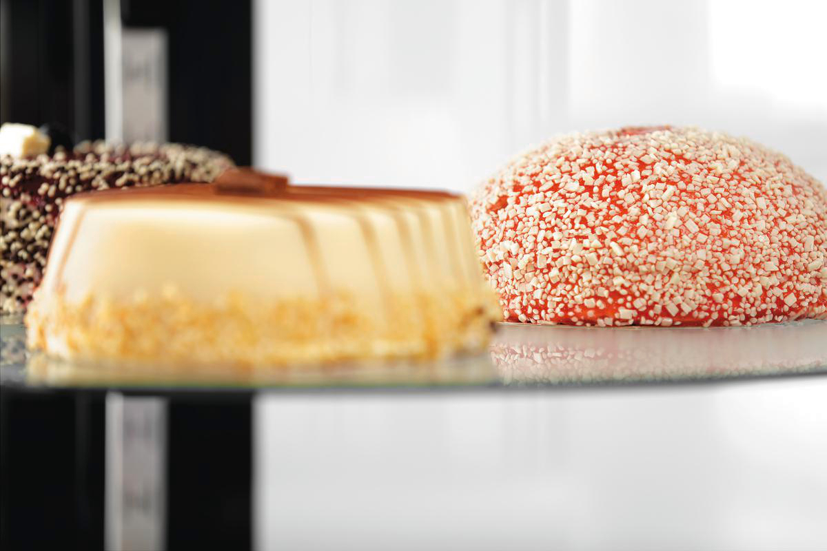 Drei verschiedene Desserts auf einer Glasplatte im Regal, darunter ein roter Kuchen mit weißen Streuseln.