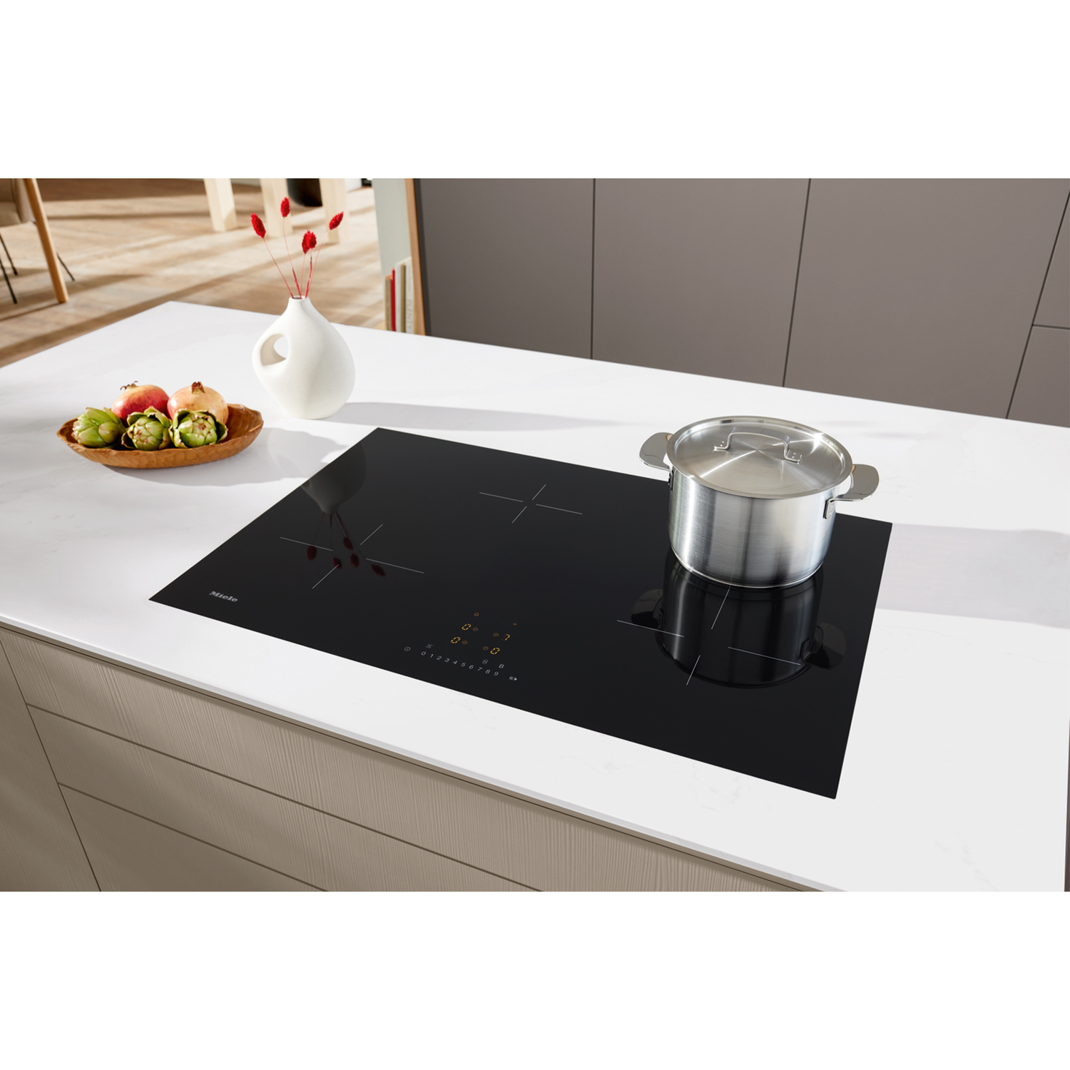 Piano cottura nero con pentola, vaso bianco con fiori rossi e vassoio di frutta.