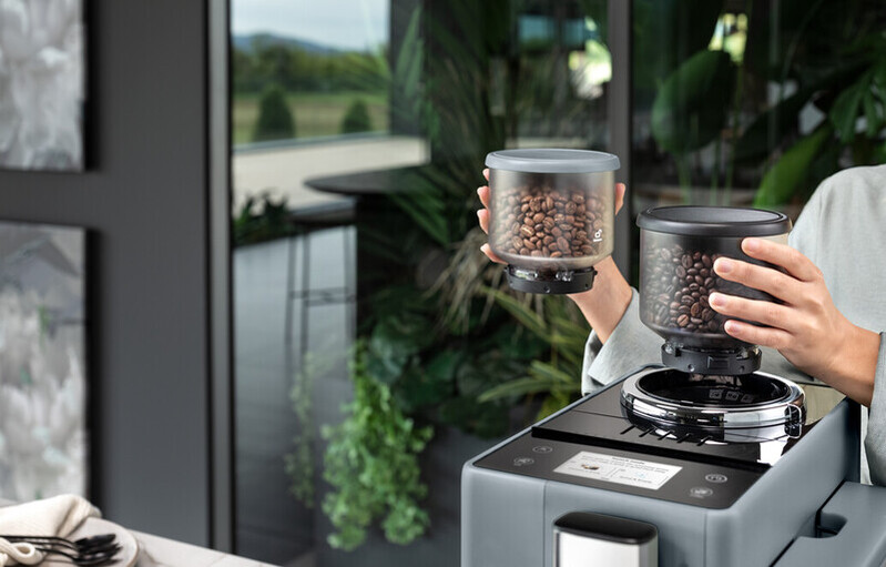 A person holding two containers of coffee beans with a coffee machine in sight.