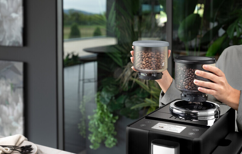 A person holding containers of coffee beans next to a coffee machine.