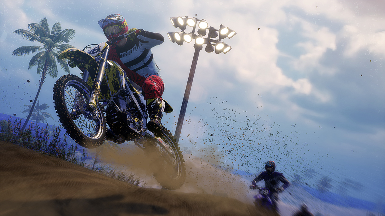 Un pilota di motocross salta su una pista sterrata, un altro lo segue. Palme sullo sfondo.