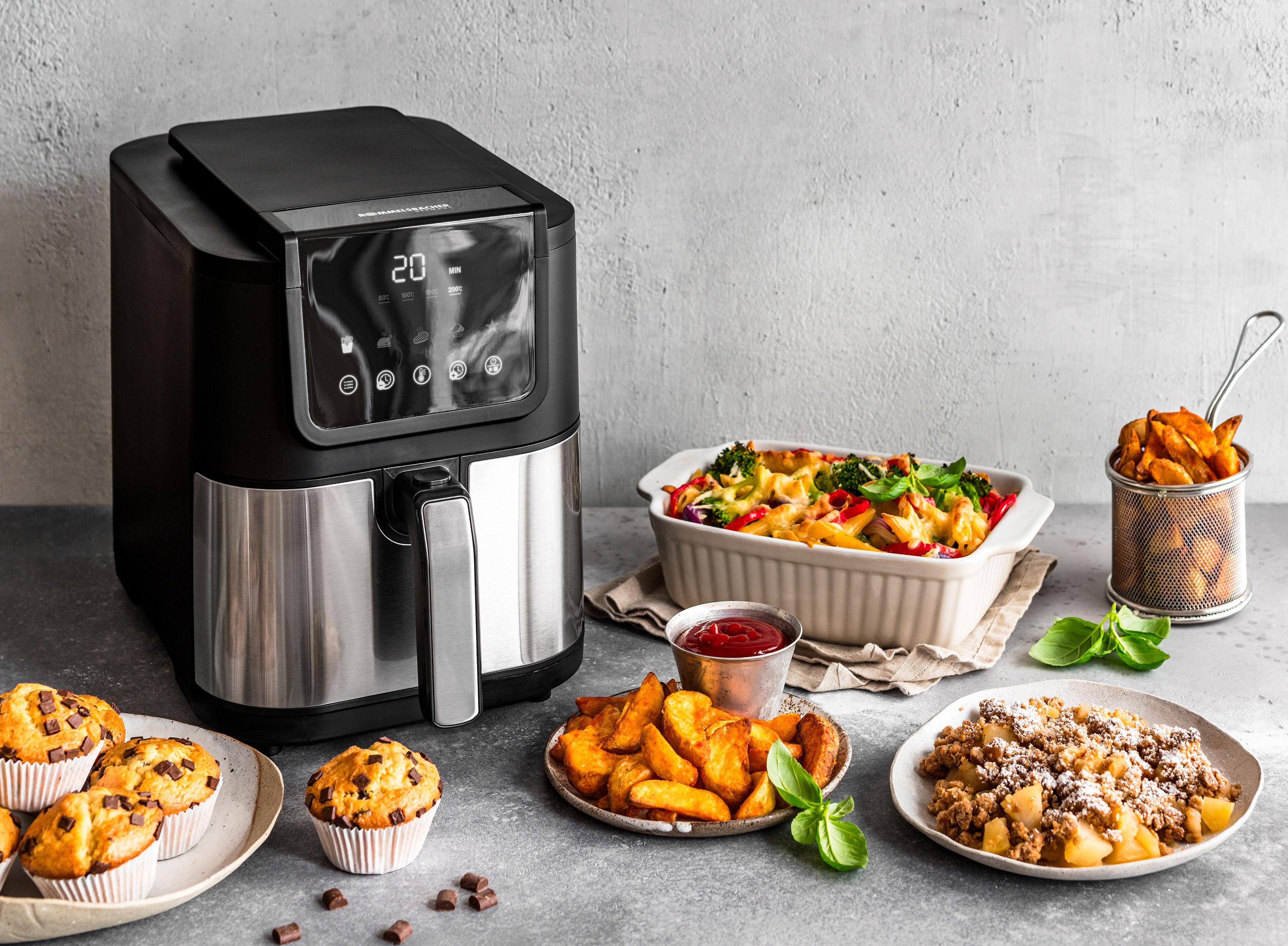 Heißluftfritteuse mit verschiedenen gekochten Speisen wie Muffins, Pommes und Gemüse auf einem Tisch.