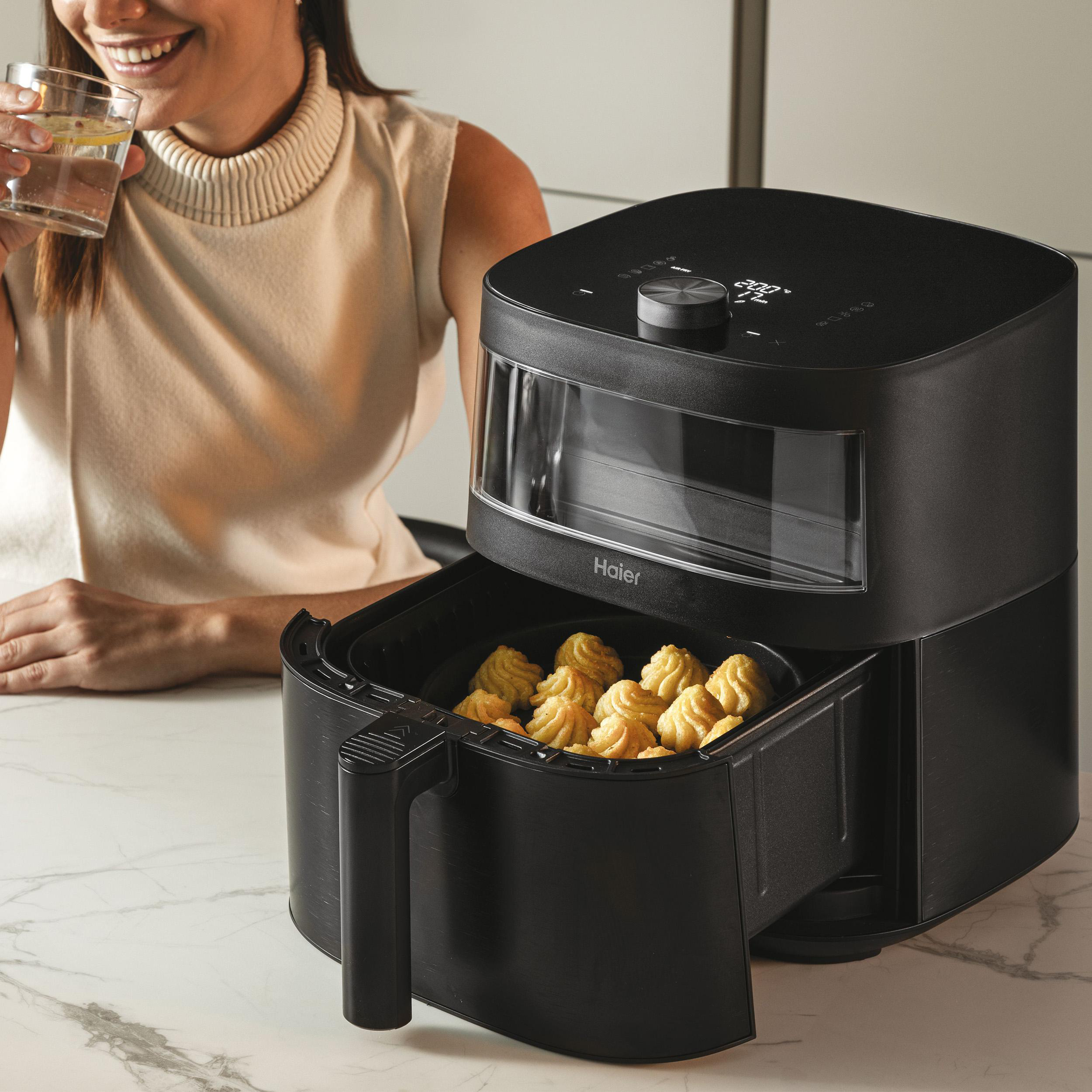 Eine Frau lächelt und hält ein Wasserglas. Eine schwarze Heißluftfritteuse mit Essen ist sichtbar.