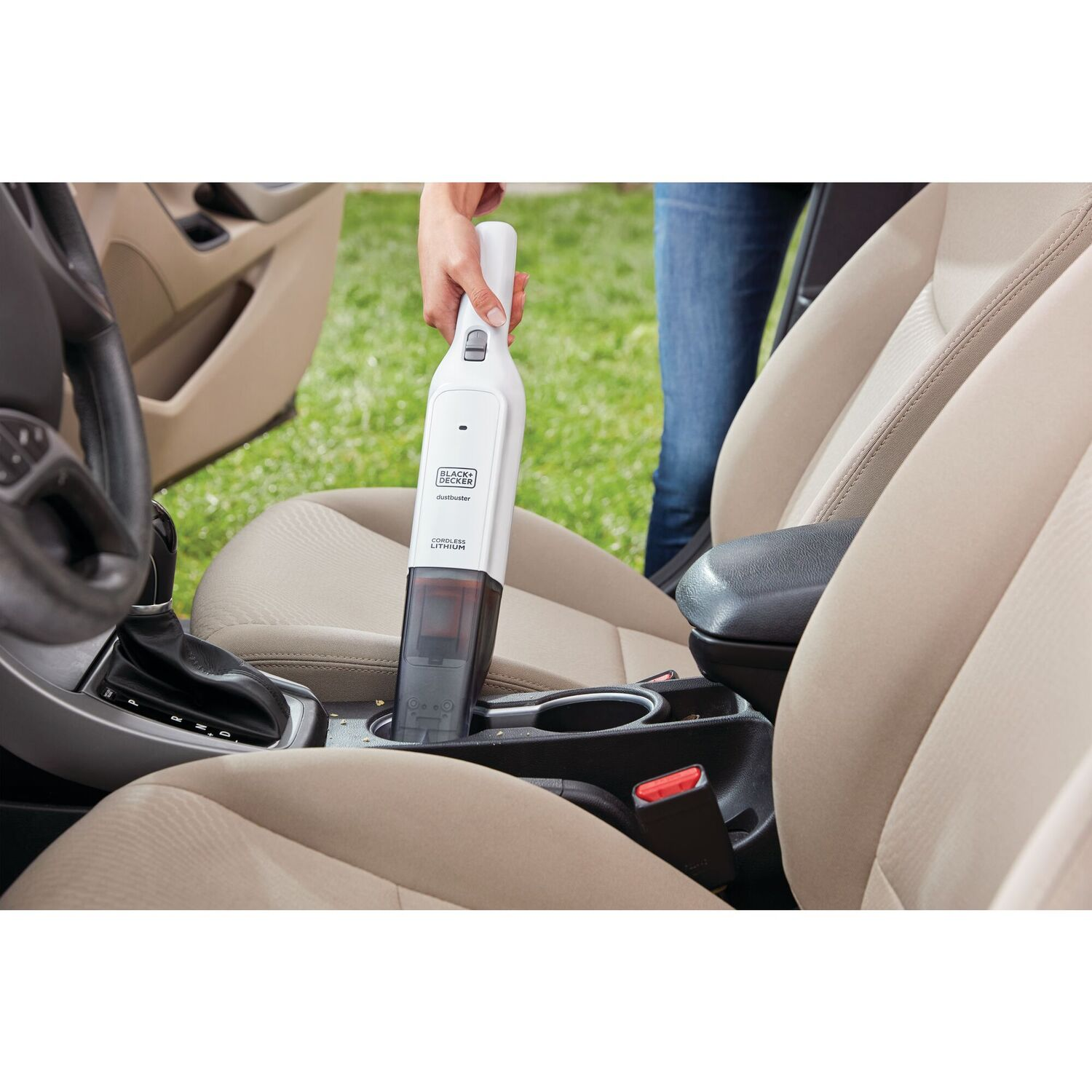 Person using a white and black cordless vacuum in a car. Beige interior with cup holder.